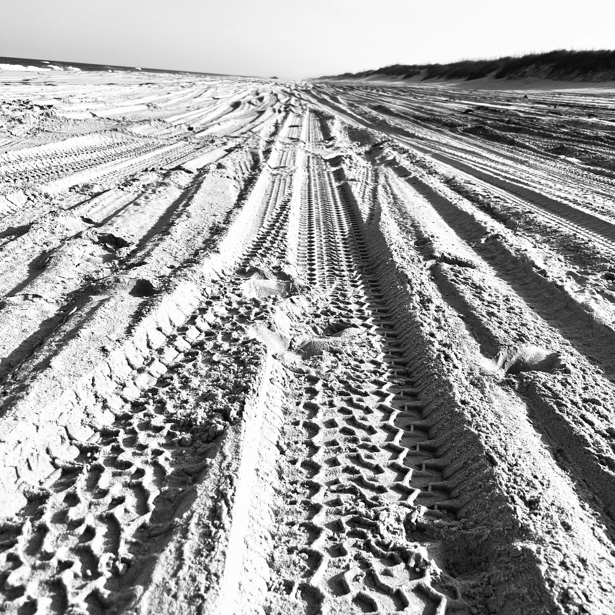 Sandy beach with tire tracks and footprints, dunes in the background under clear sky