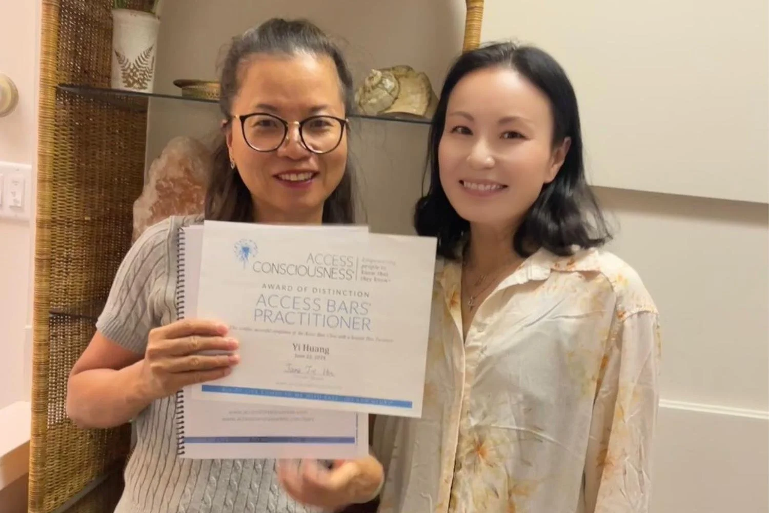Two women standing together indoors, one holding a certificate that reads 'Access Bars Practitioner'. They are smiling at the camera, one with glasses and the other with black hair, in casual attire.