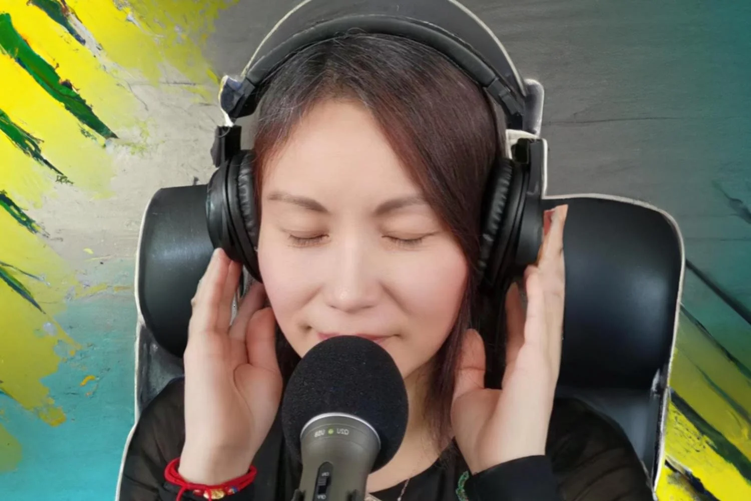 Young woman with closed eyes wearing over-ear headphones, holding them with both hands, sitting in front of a microphone in a colorful studio setting.