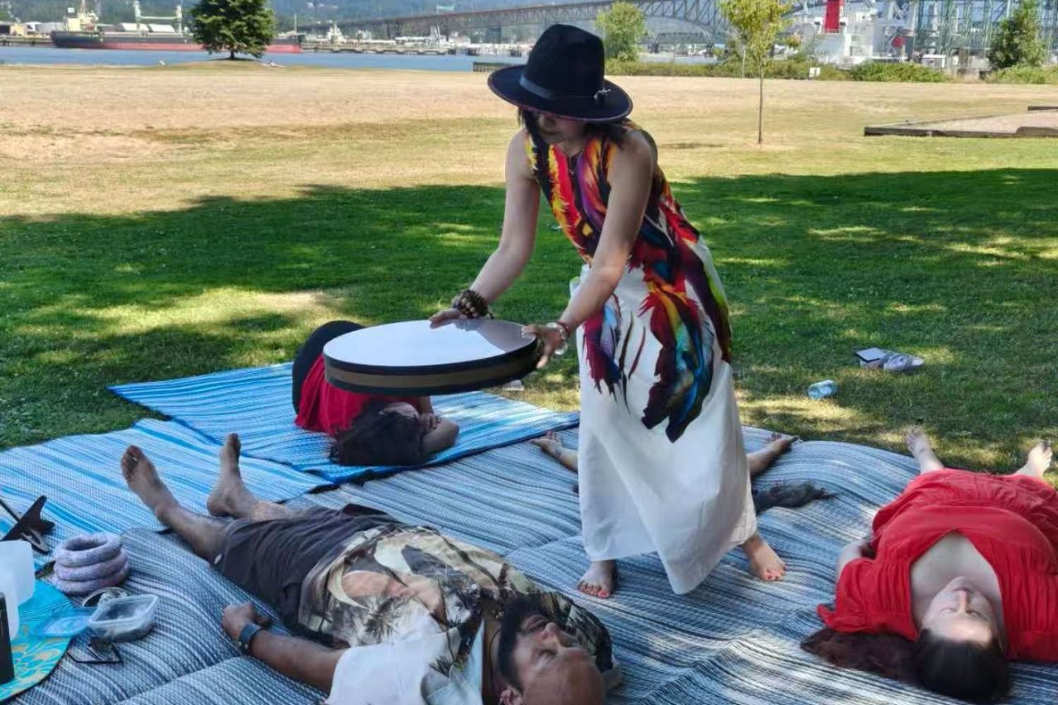 People relaxing on blankets outdoors near a river, with one person lying down and two women standing, one with a black hat and colorful dress, and the other in a red dress. The woman with the hat is holding a circular object over one person lying on the ground.
