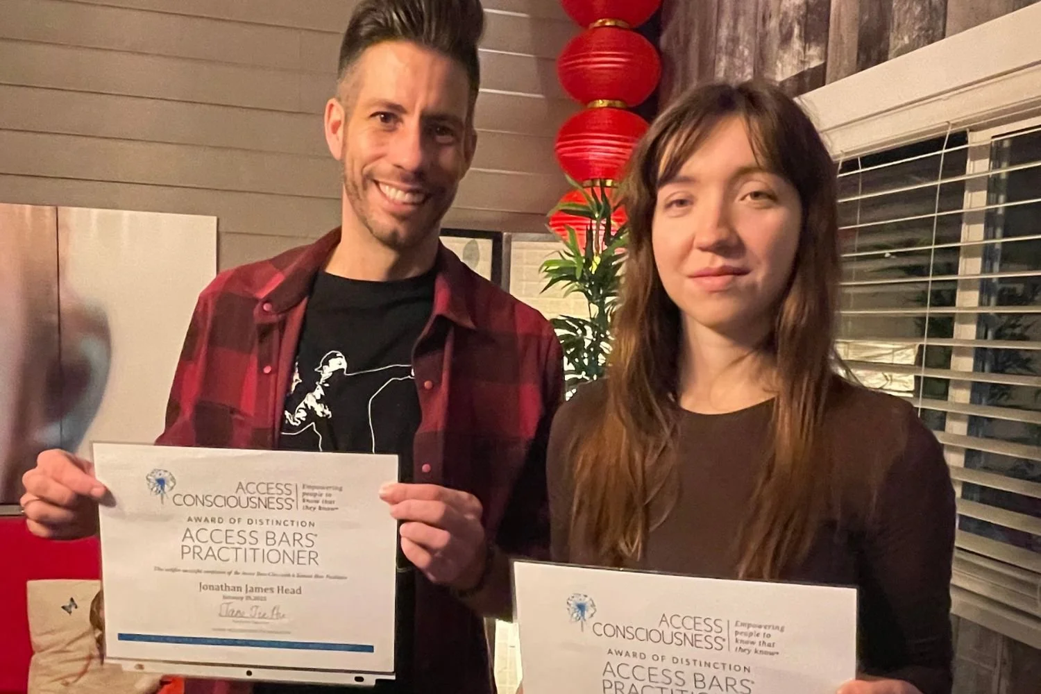 A man and a woman holding certificates, smiling, indoors with red lanterns, a plant, and blinds behind them.