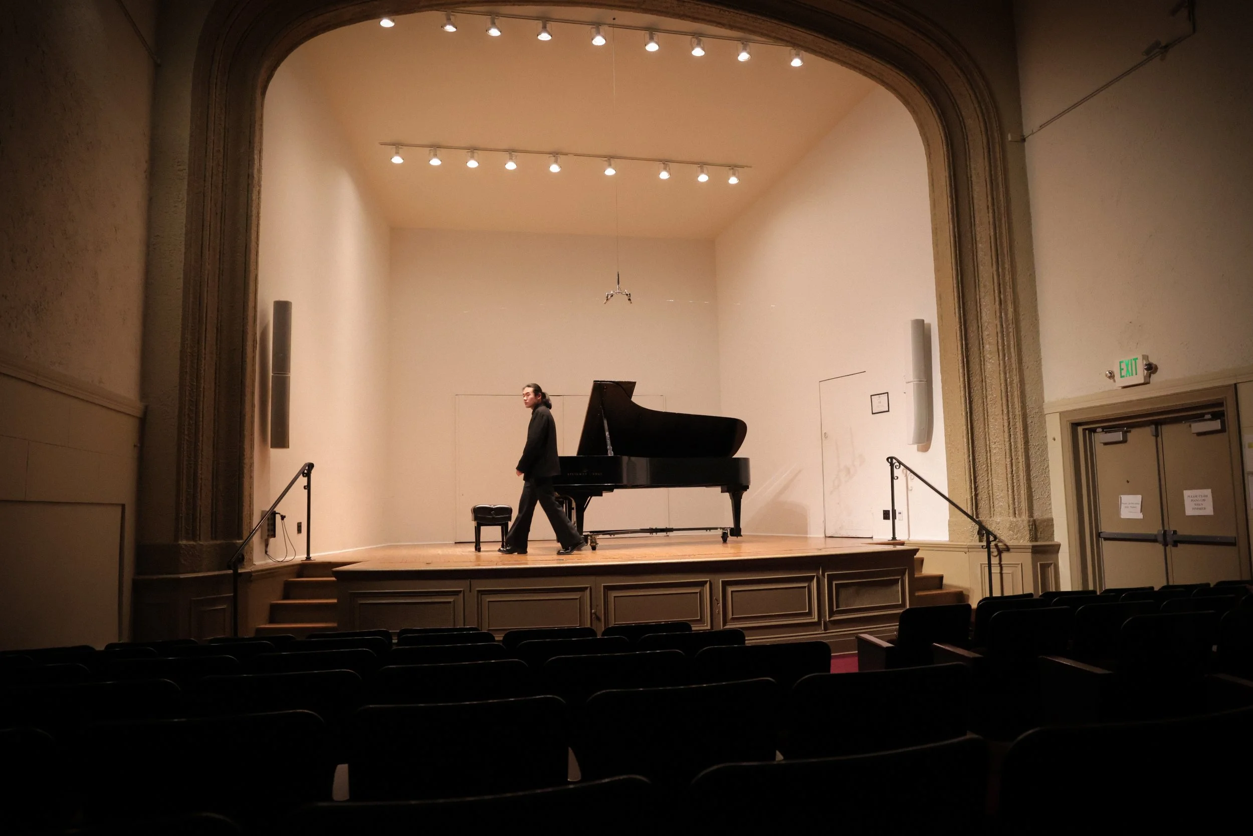 A person walking on a stage with a grand piano, in an empty auditorium with rows of seats and a high ceiling with spotlights.