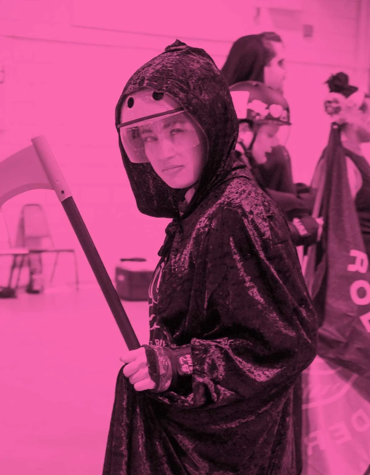 A person in a velvety black cloack and transparent face shield holding a scyth, standing indoors with pink-tinted lighting, other people and derby gear in the background.