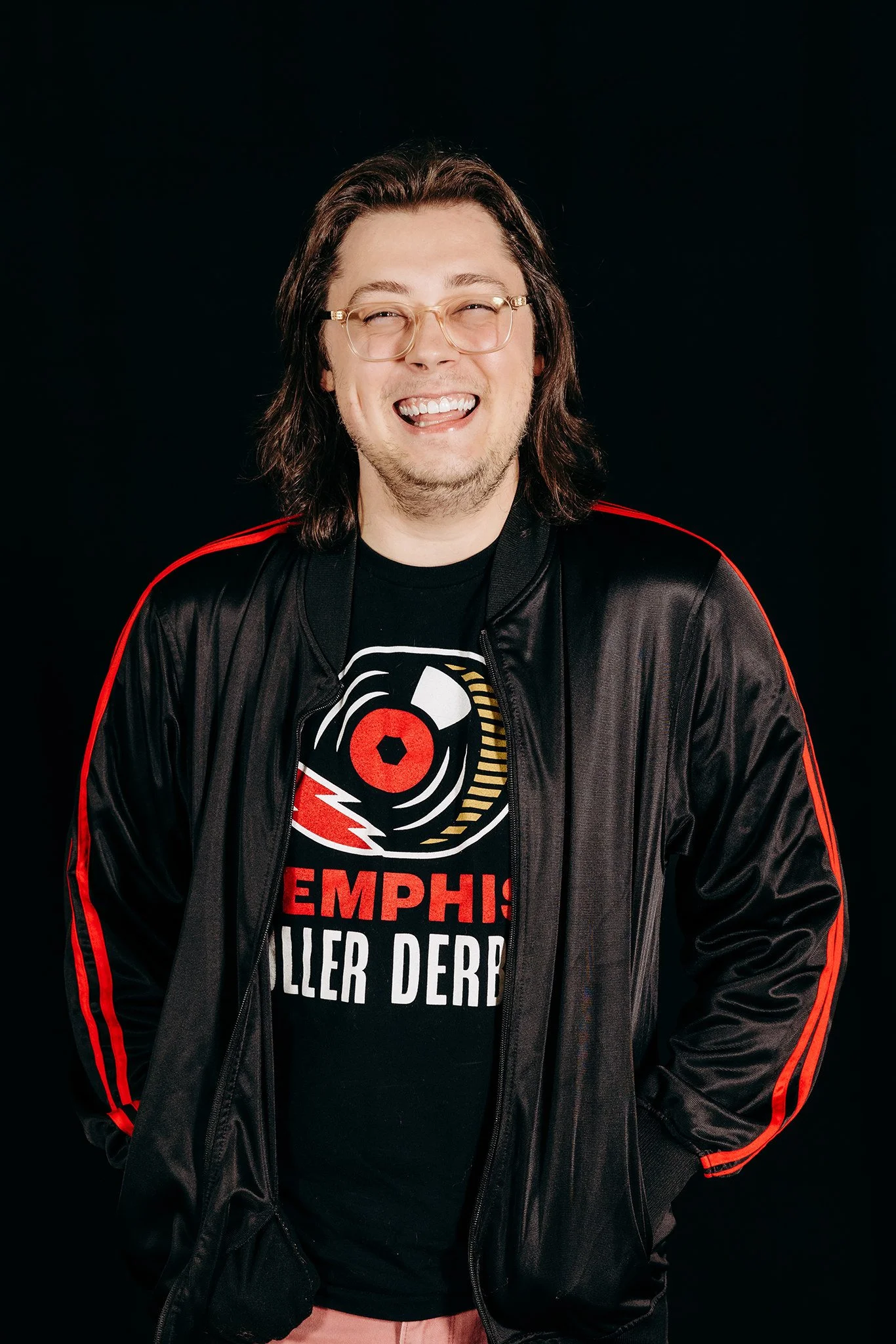 A man with shoulder-length brown hair, glasses, and a beard, smiling widely, wearing a black Memphis Roller Derby t-shirt and a black jacket with red stripes on the sleeves, standing against a black background.