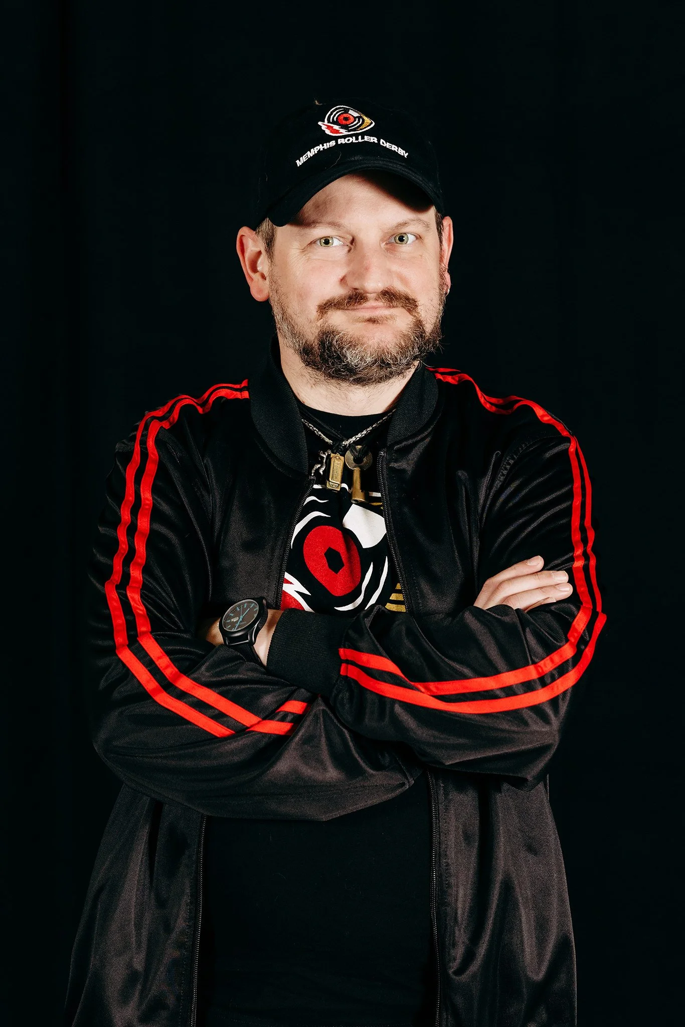 Man wearing a black cap with Memphis Roller Derby patch, black jacket with red stripes, and crossed arms, standing against a black background.