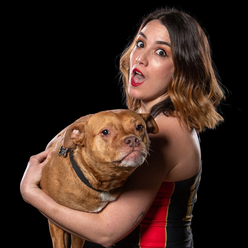 A woman with shoulder-length wavy brown hair, wearing a sleeveless black top with red and yellow stripes, holding a brown dog with a black collar, both against a black background. The woman appears surprised or excited, with wide eyes and open mouth.