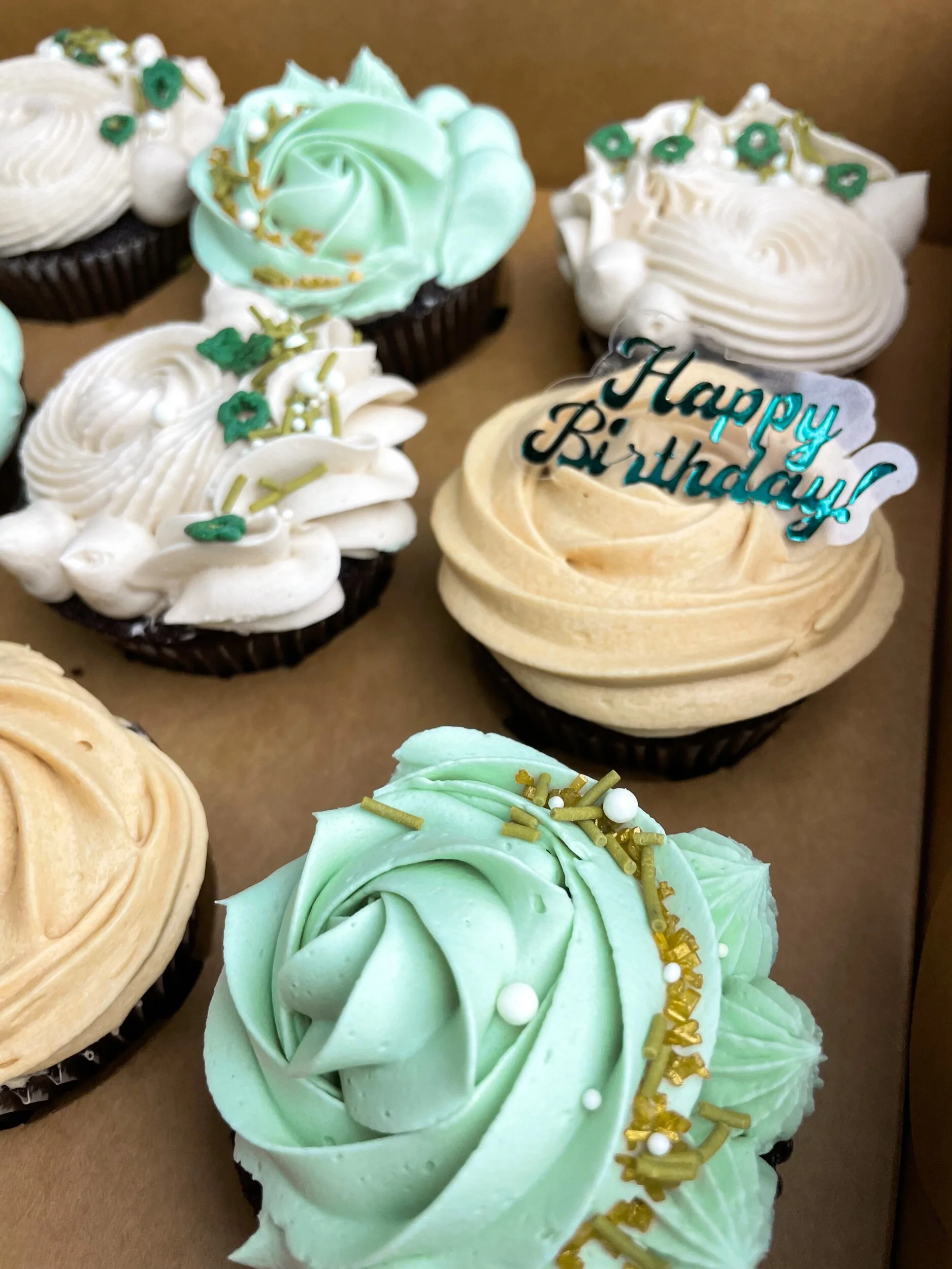 Assorted frosted cupcakes in a box, featuring pastel colors and decorated with sprinkles and sugar embellishments. One cupcake has a "Happy Birthday" topper.