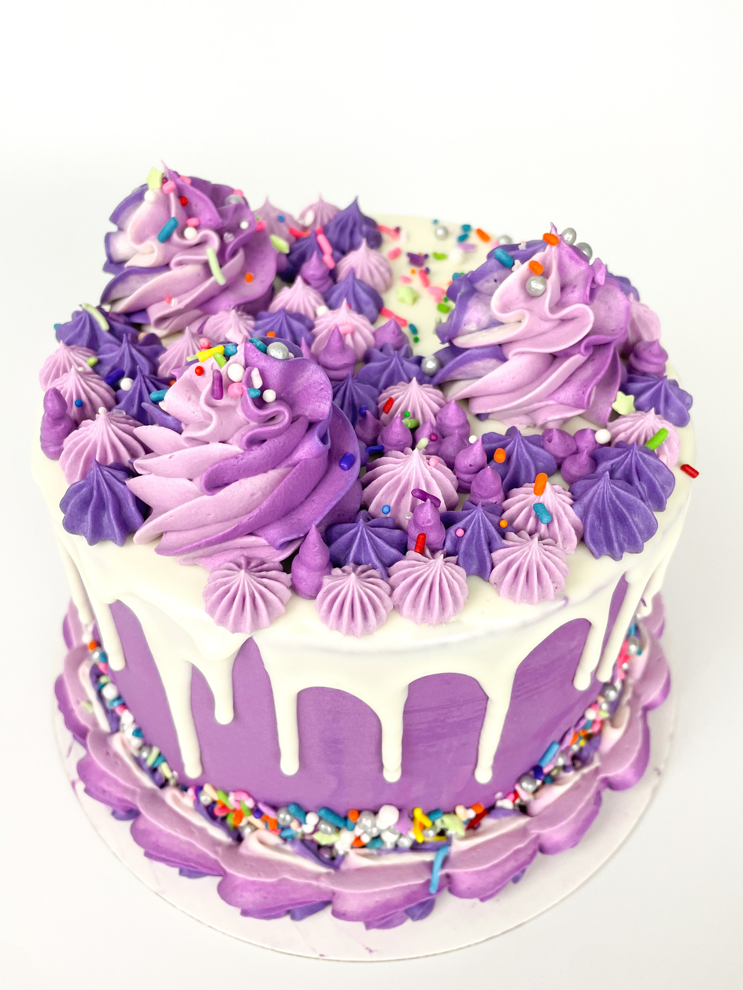 Purple frosted cake with white drip and colorful sprinkles.