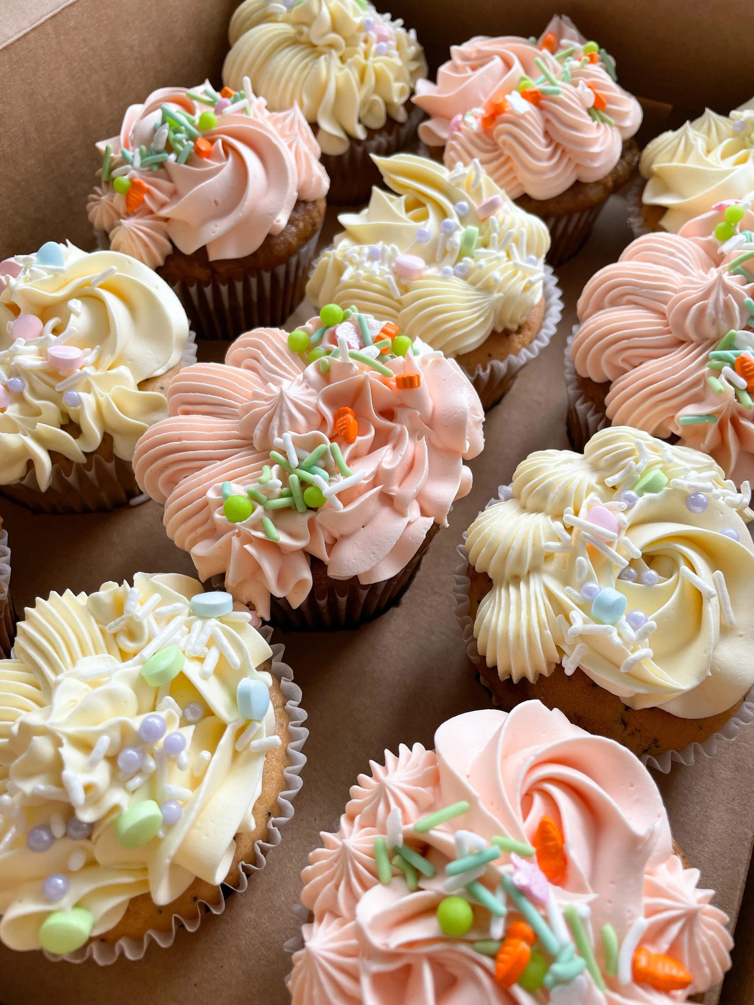 Box of cupcakes with pink and yellow frosting topped with colorful sprinkles.