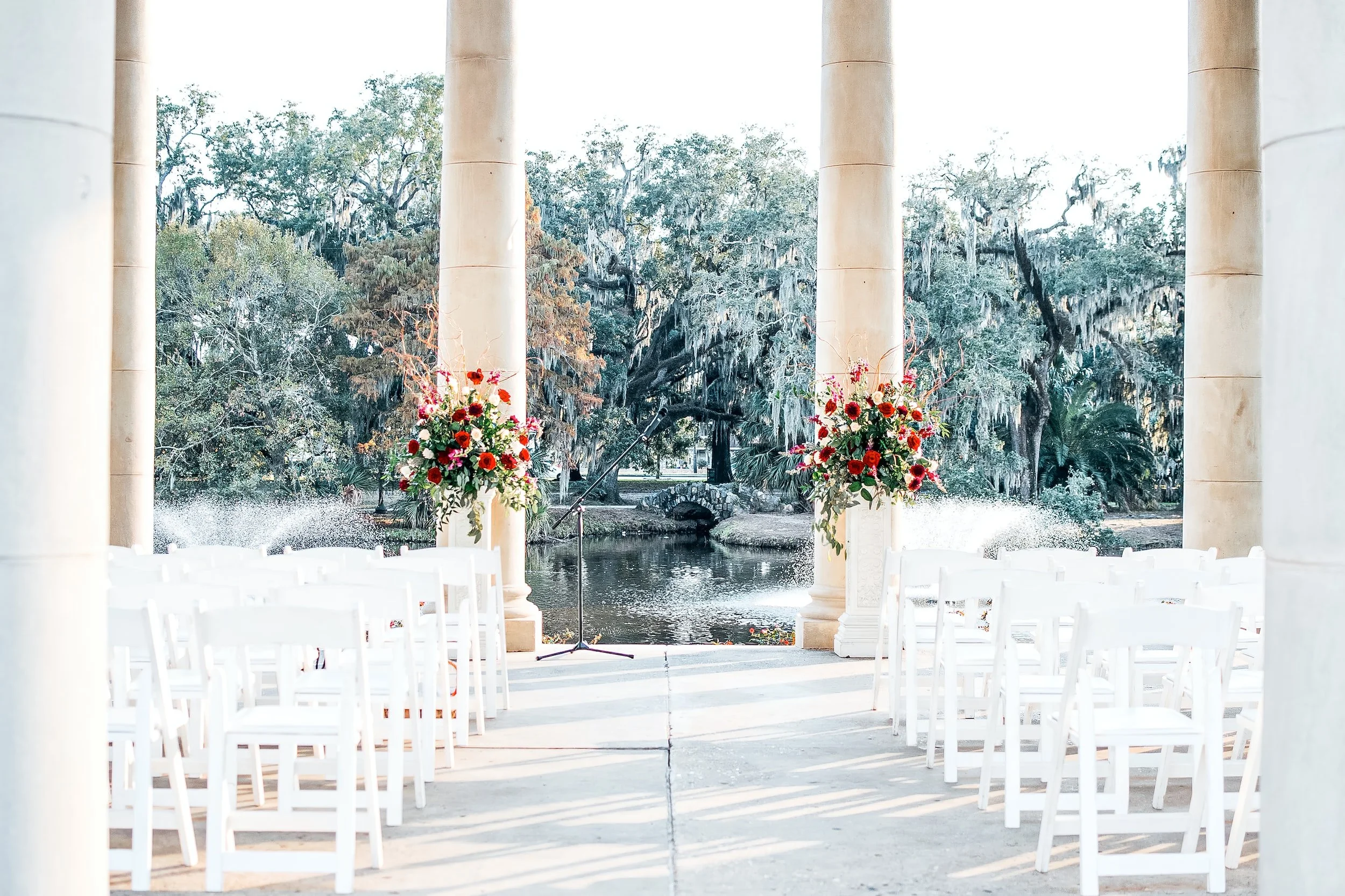 Outdoor wedding ceremony setup with white chairs, floral arrangements, and a pond framed by large columns in a garden