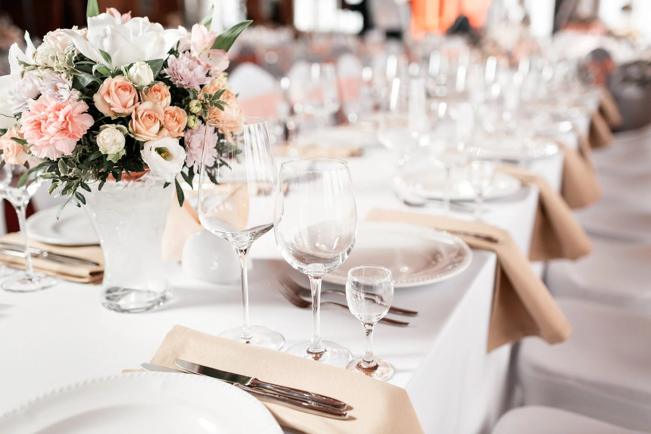 Elegant banquet table setting with a large floral centerpiece of pink and white roses, carnations, and greenery, with white plates, silverware on beige napkins, and multiple wine glasses arranged on a white tablecloth.
