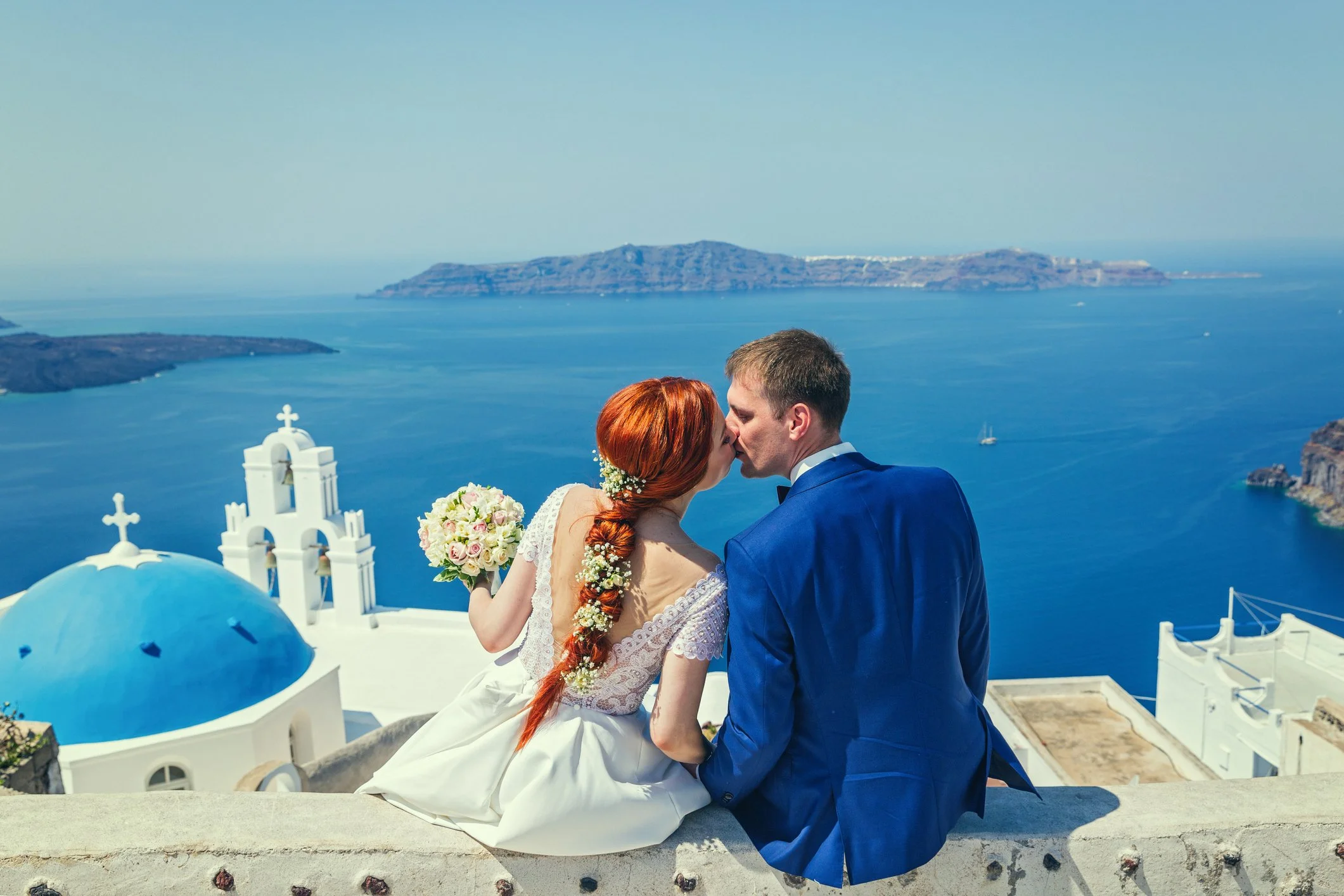 A newlywed couple kissing with a scenic Greek island view, white buildings with blue domes, against the Aegean Sea.
