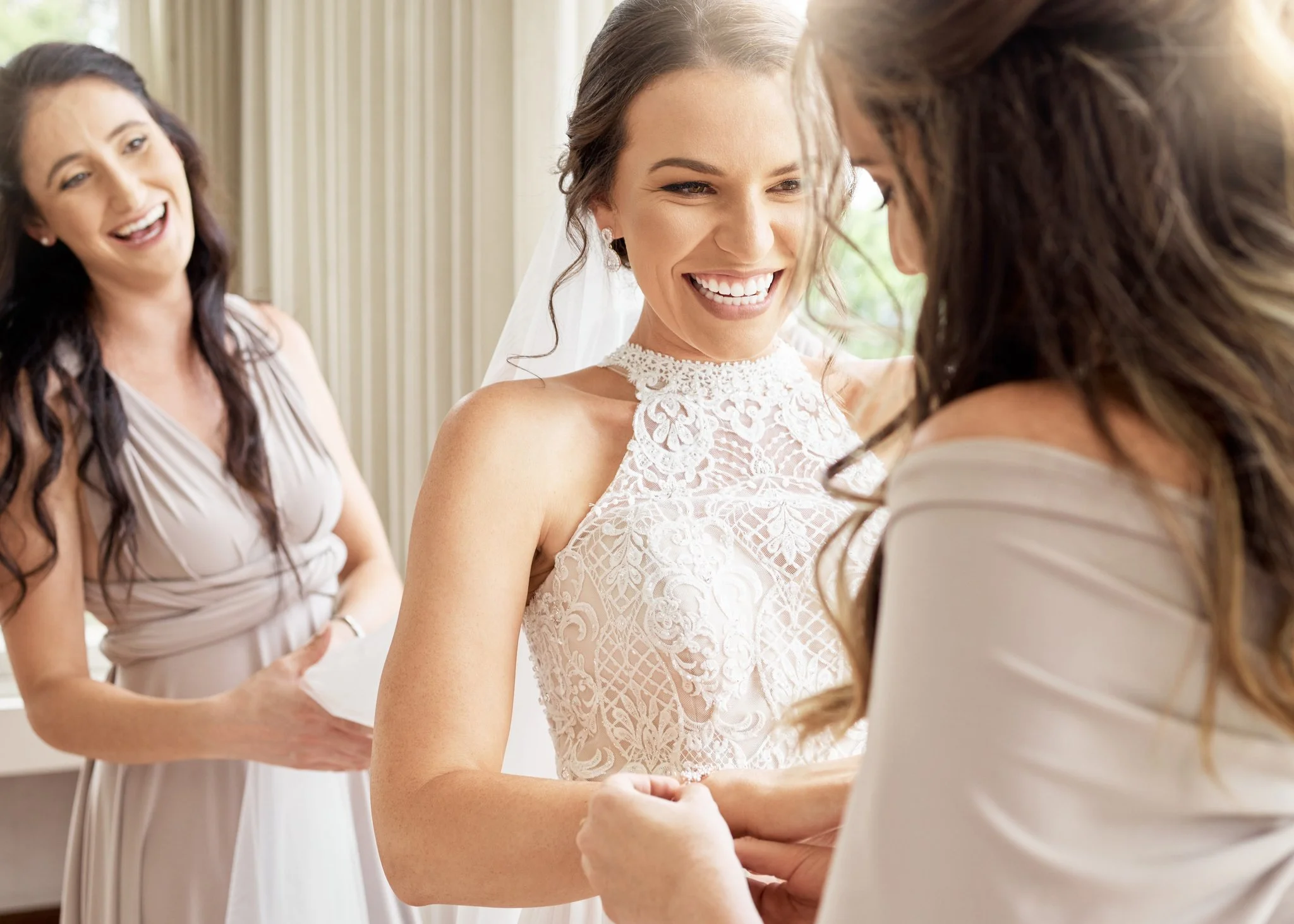 Bride smiling as she is helped into her wedding dress by her bridesmaids, with another bridesmaid smiling in the background.