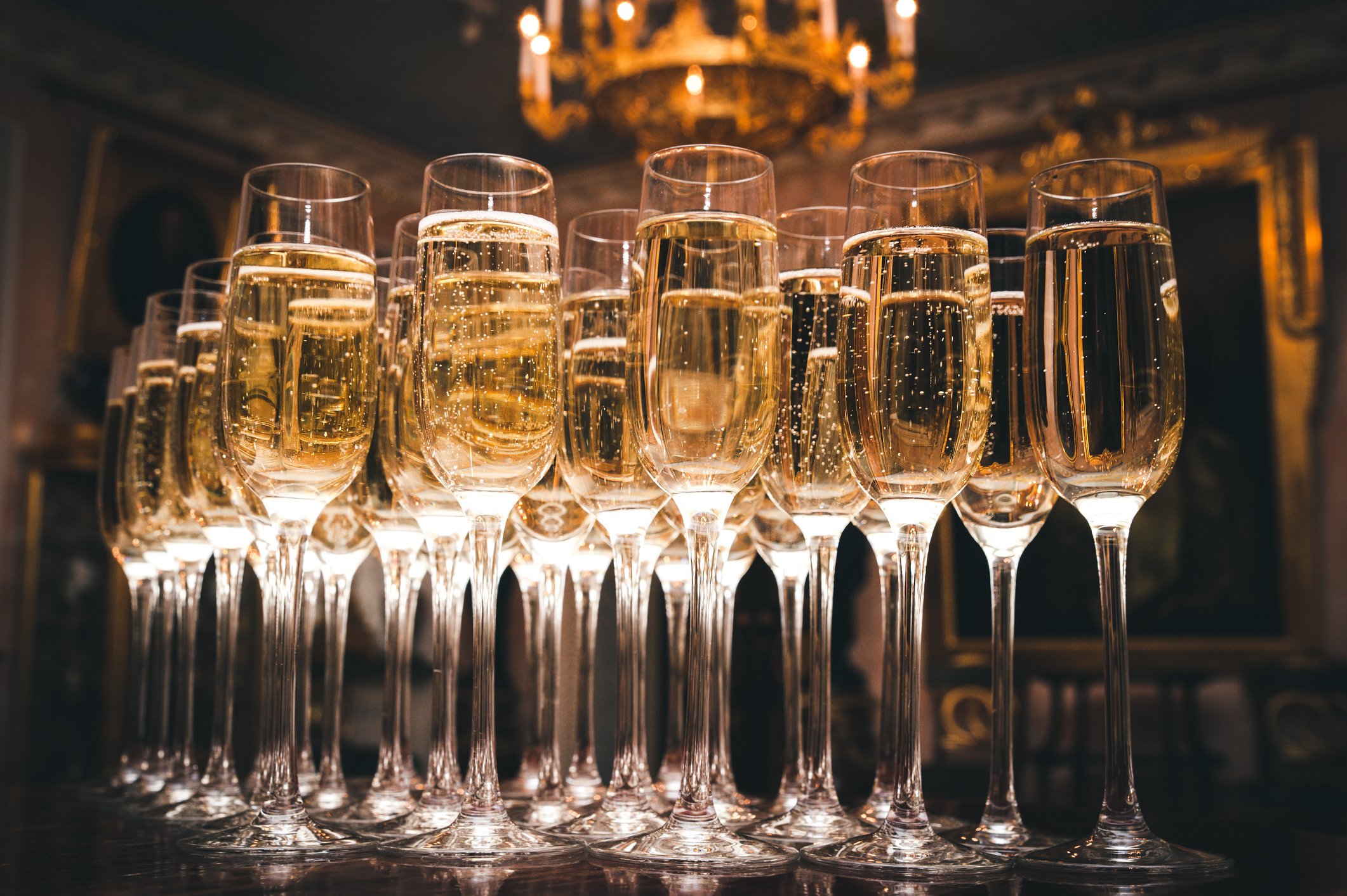 Multiple filled champagne flutes arranged on a table with a chandelier overhead in an elegant, dimly-lit room.