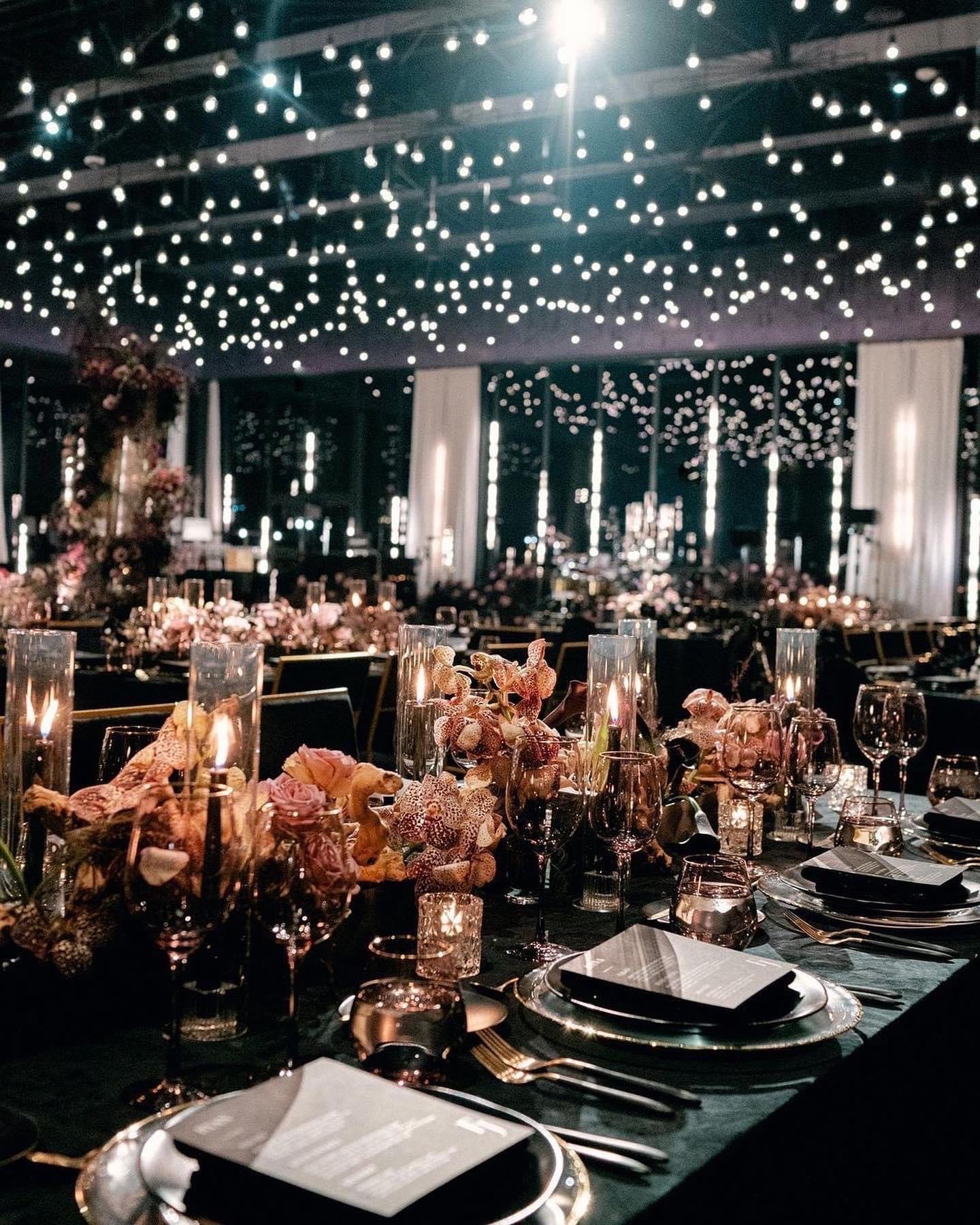 Elegant event setup with a long table decorated with flower arrangements, candles in glass holders, and fine china in a dimly lit room with a string of lights overhead.