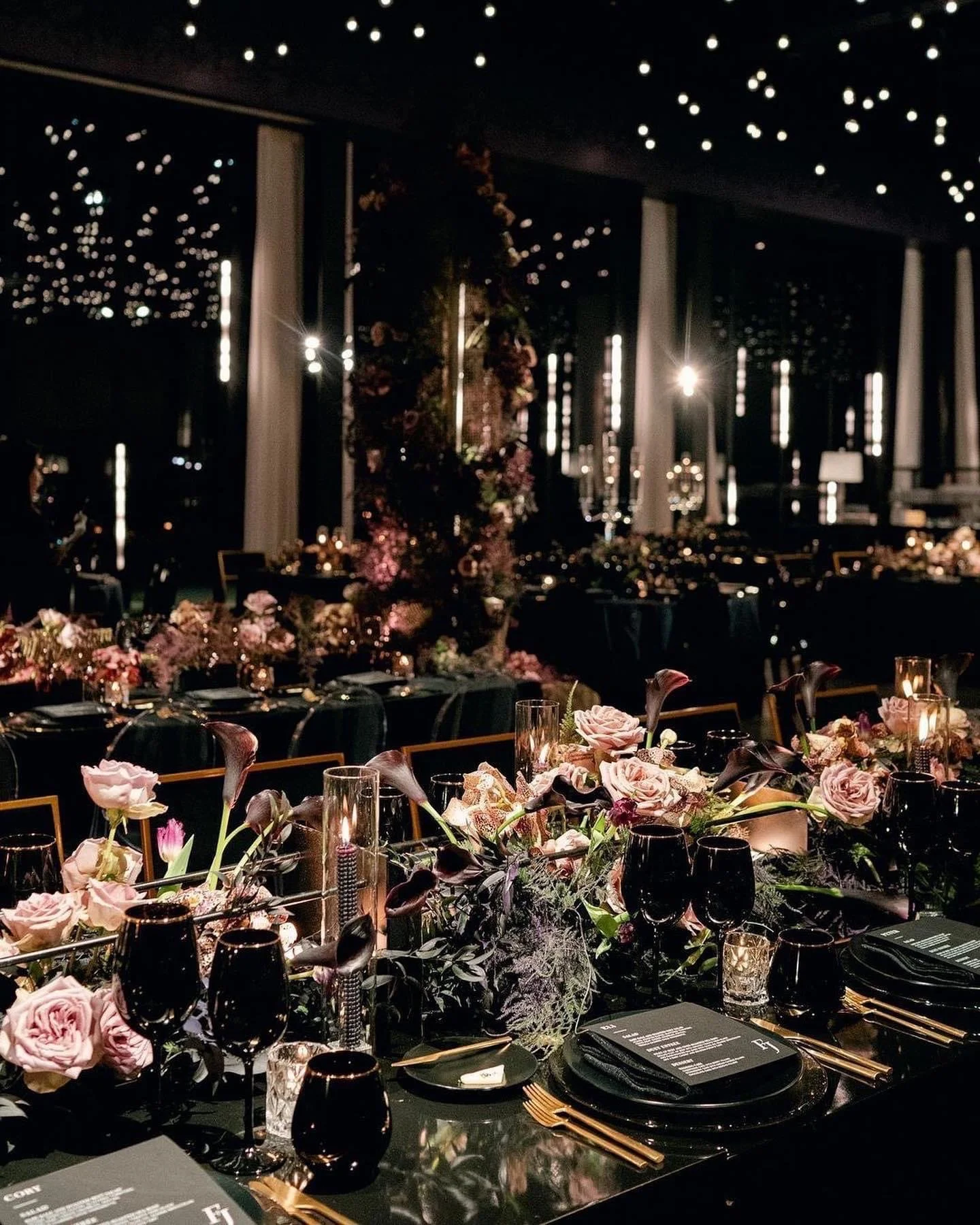Elegant banquet table decorated with pink and purple flowers, candles, black glassware, and gold cutlery in a dimly lit event space with ceiling star lights and floral centerpieces.