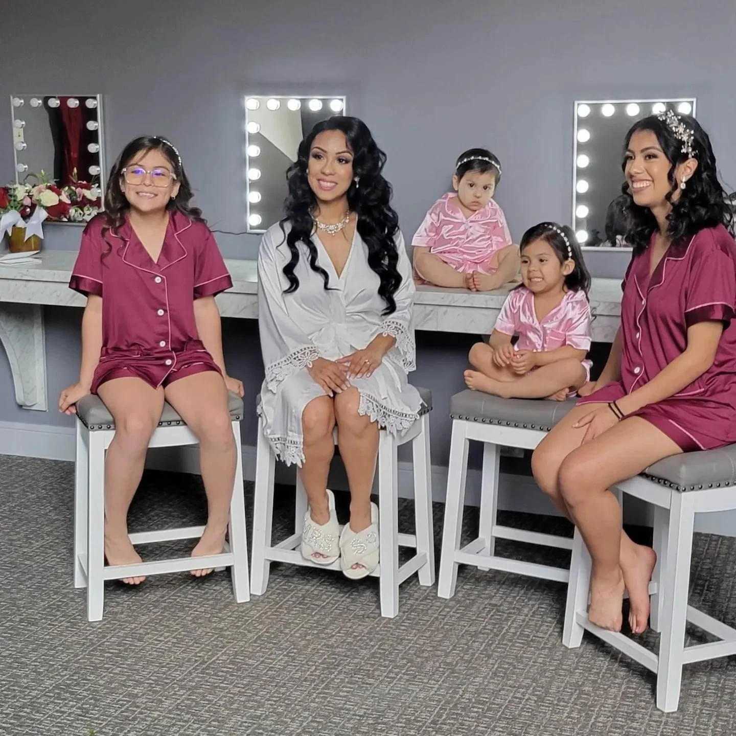 A woman and four young girls sitting on stools in a dressing room with mirrors surrounded by light bulbs, all wearing matching pink satin pajamas, and the woman in a white satin robe, all smiling.