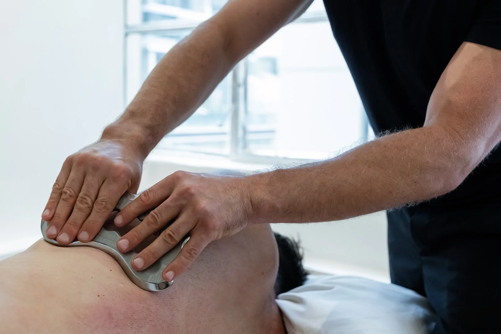 Close-up view of a therapist performing remedial massage therapy using a metal tool on a client's back, focusing on deep tissue treatment to relieve muscle tension and improve circulation.