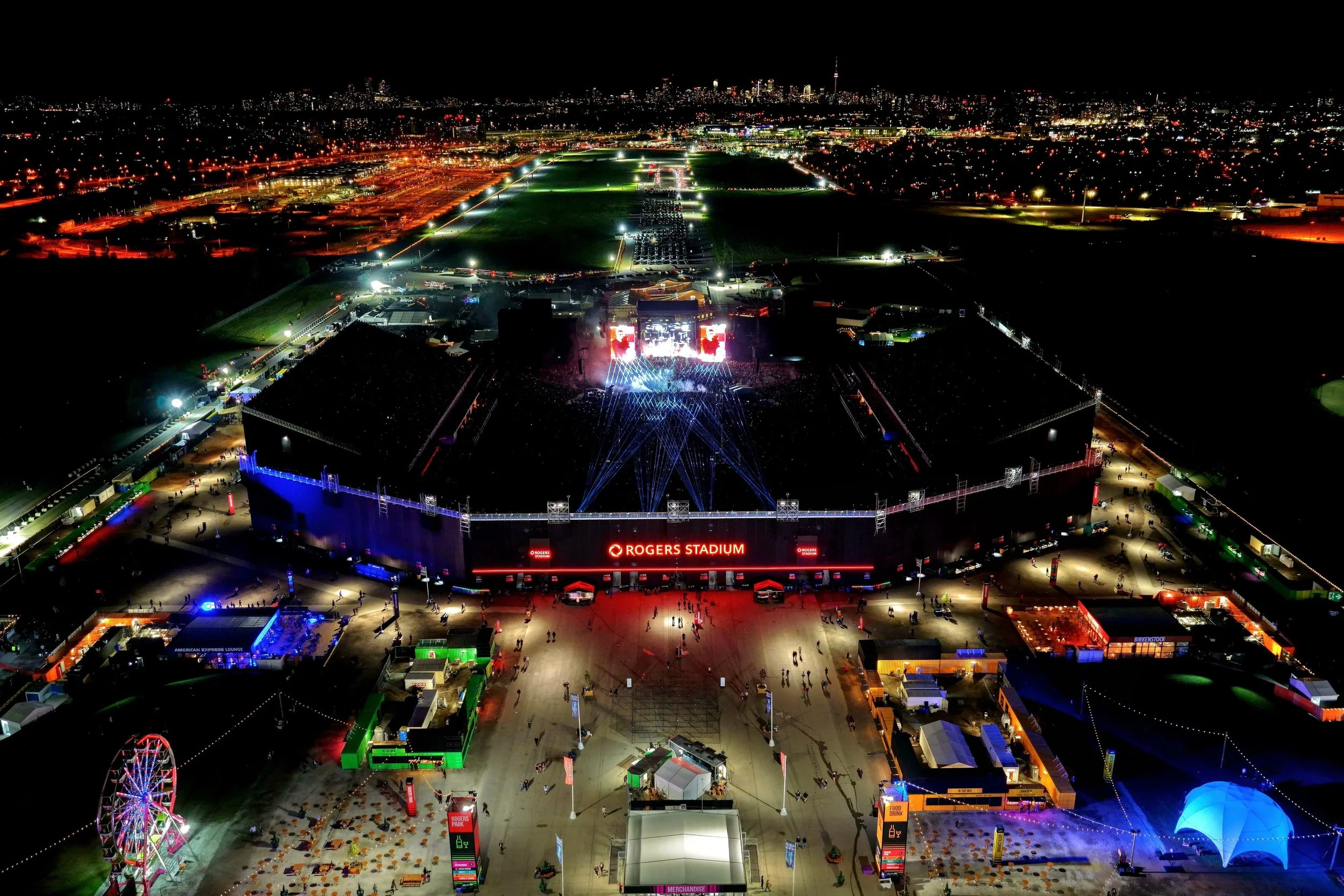 Nighttime aerial view of a large outdoor concert with bright lights, a stage, and a crowd in front, with a city skyline in the background.