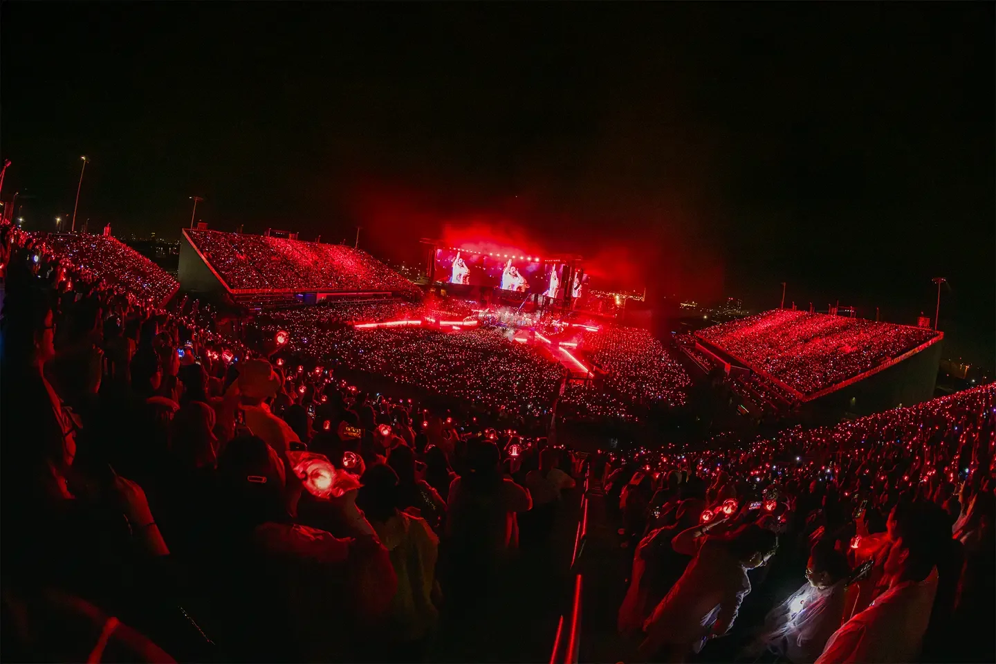 Large outdoor concert at night with a stage, big screens, and a crowd illuminated by red lights, creating a vibrant atmosphere.