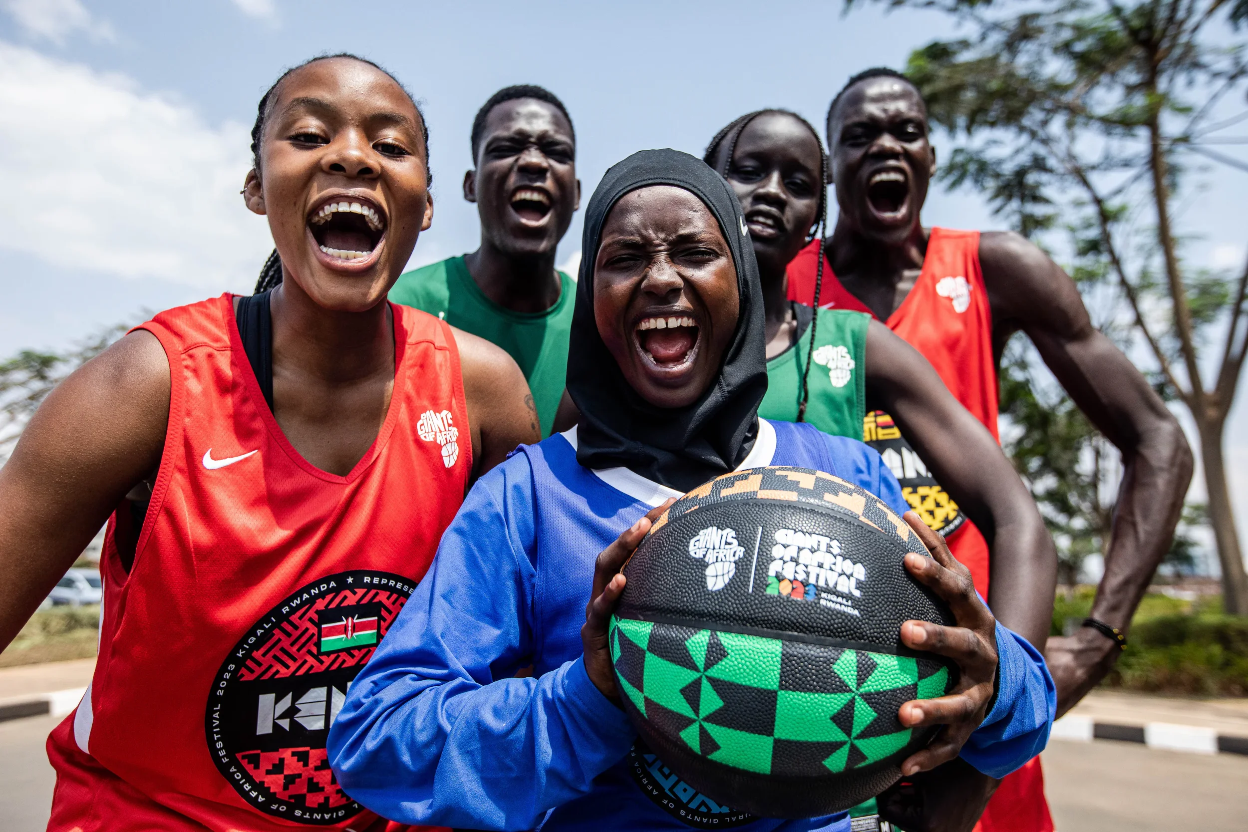 Group of smiling athletes, four women and one man, standing outdoors with trees in the background, holding a basketball with a colorful design, celebrating at a sports event.