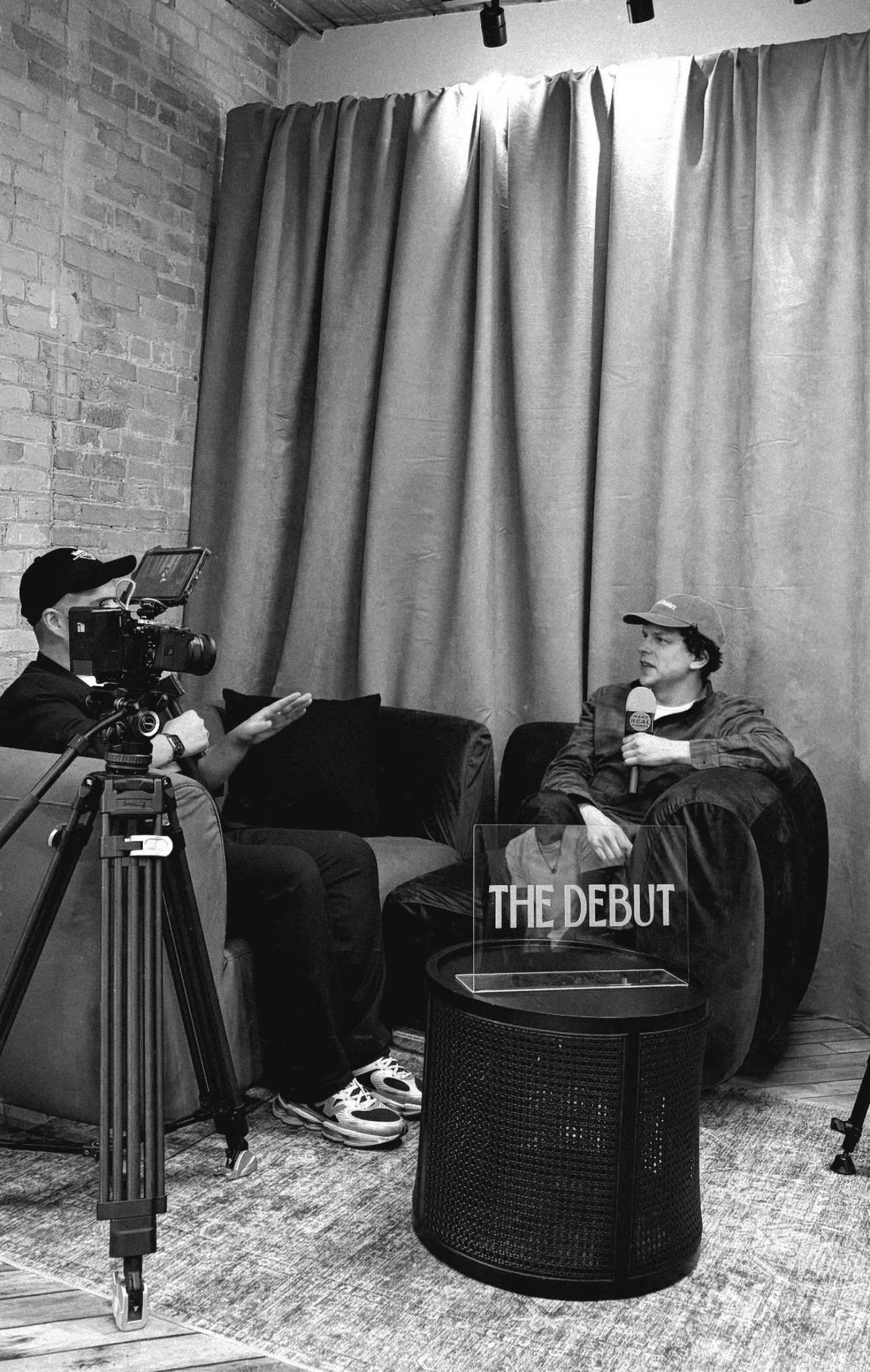 A man with curly hair and a cap is talking into a microphone during an interview while another person is operating a camera. There is a sign on a table in front of them that reads 'The Debut'. The scene is indoors, with a brick wall and large curtains in the background.