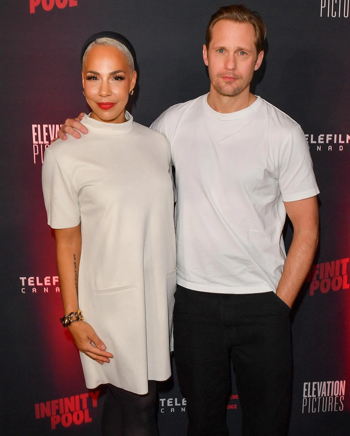 A woman and a man standing together in front of a black backdrop with red and white text. The woman is wearing a white dress with a high neckline, gold jewelry, and has short, platinum blonde hair. The man is wearing a white t-shirt and black pants, with light brown hair and a beard. They are both looking at the camera, with the woman's left hand resting on the man's shoulder.
