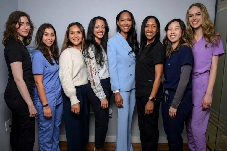 Group of eight women standing together, smiling, in healthcare uniforms and business attire, in a room with a gray wall background.