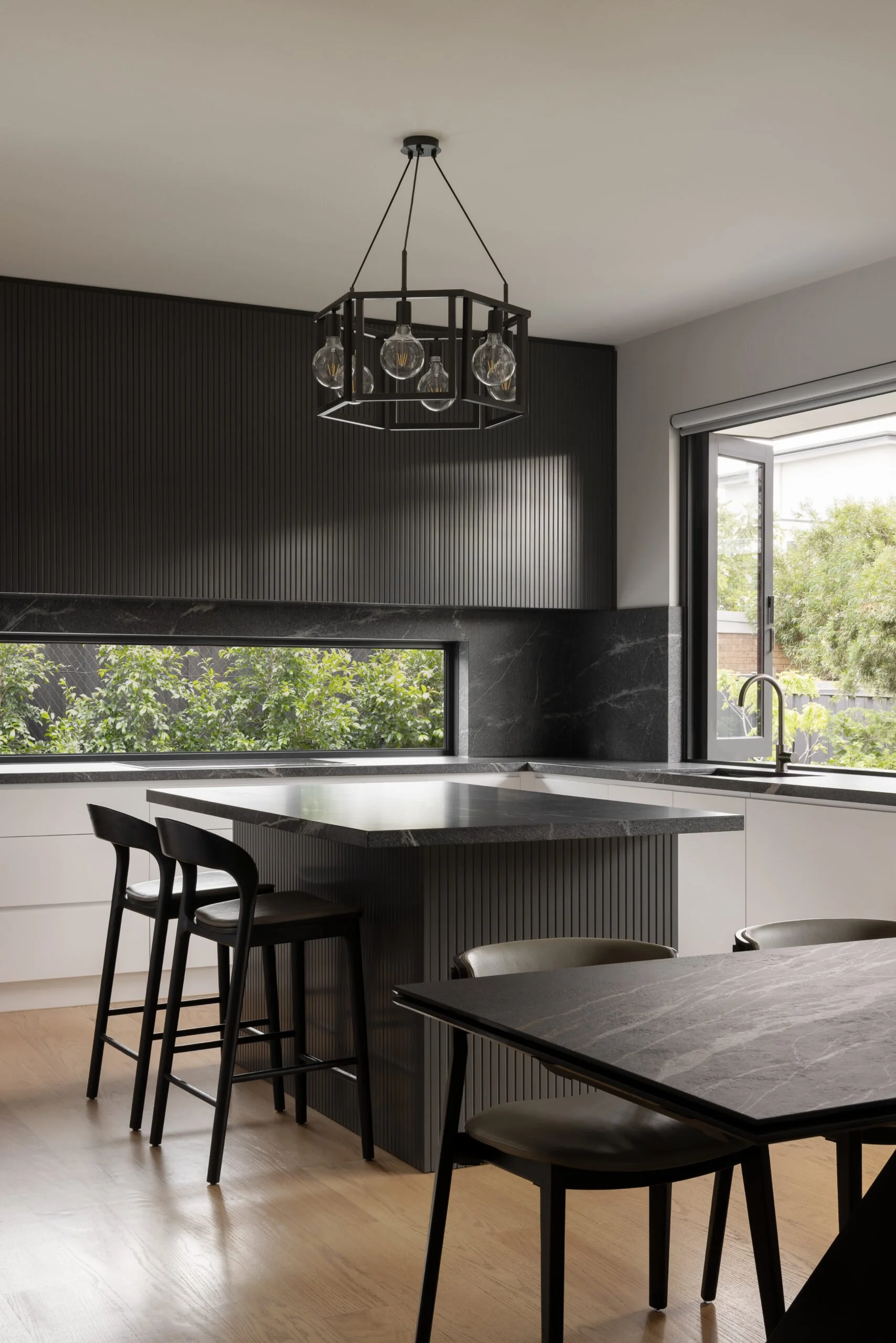 Contemporary Melbourne apartment kitchen designed for entertaining, featuring a functional island layout, integrated storage, fluted black joinery and a long picture window framing greenery.