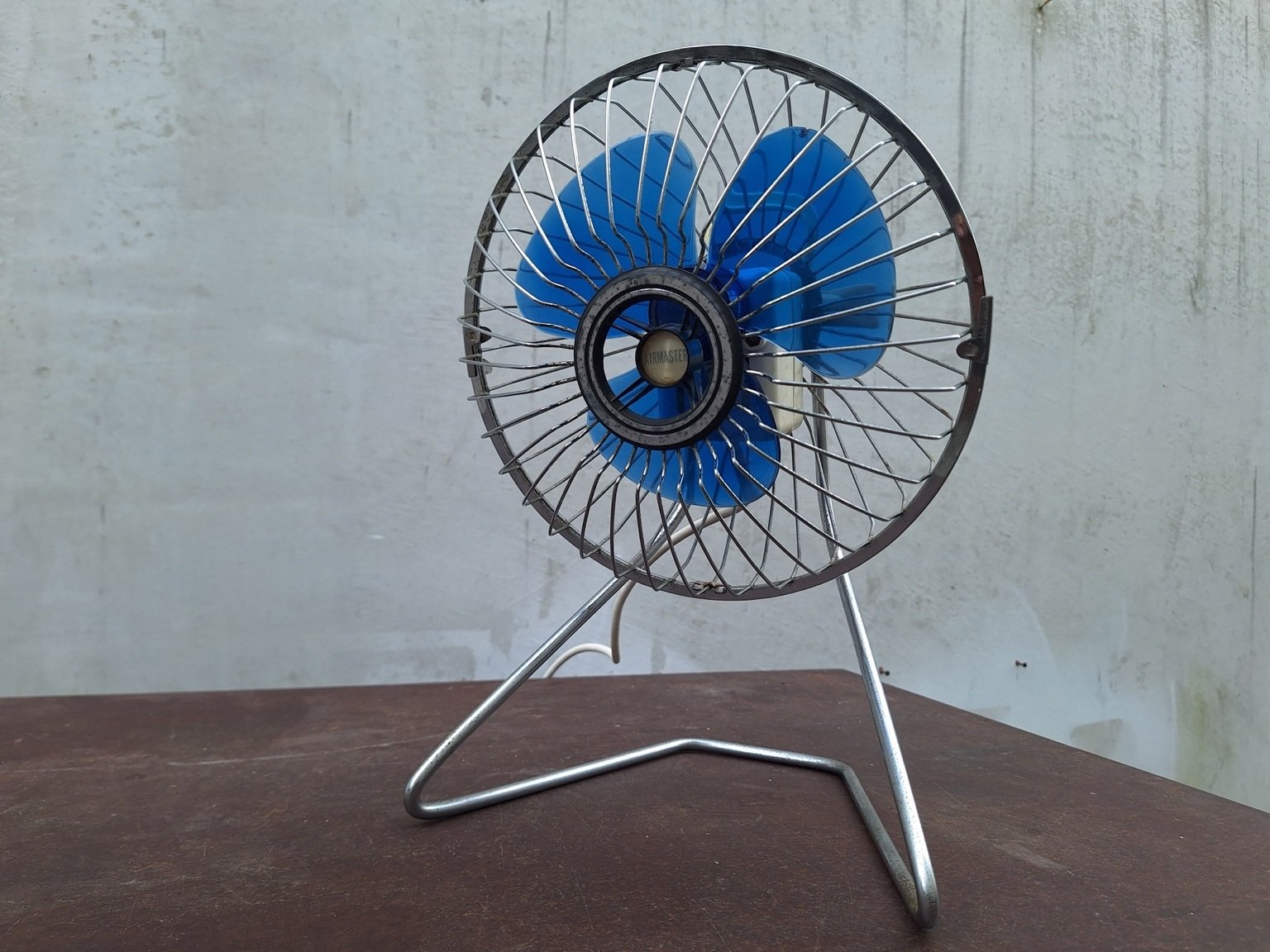 Vintage Airmaster desk fan with blue blades, circular metal grille and bent metal stand, viewed from the front at an angle.