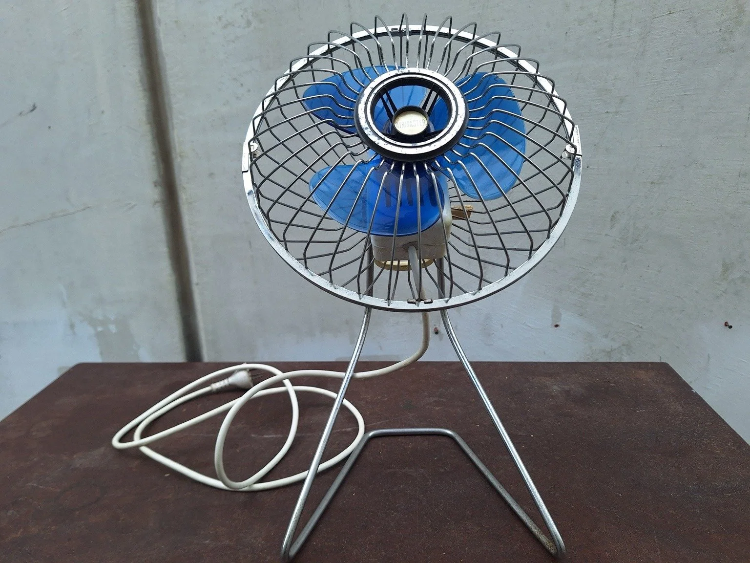 Front view of the Airmaster fan showing its circular grille, blue blades, wire stand and power cord.