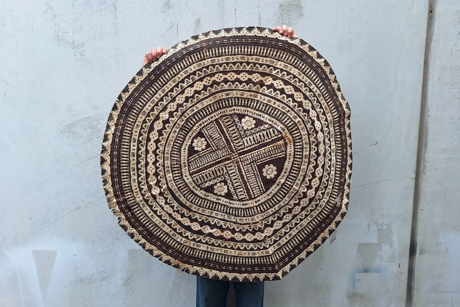 Full view of a circular tapa barkcloth with concentric cream, black and reddish-brown geometric and floral patterns.