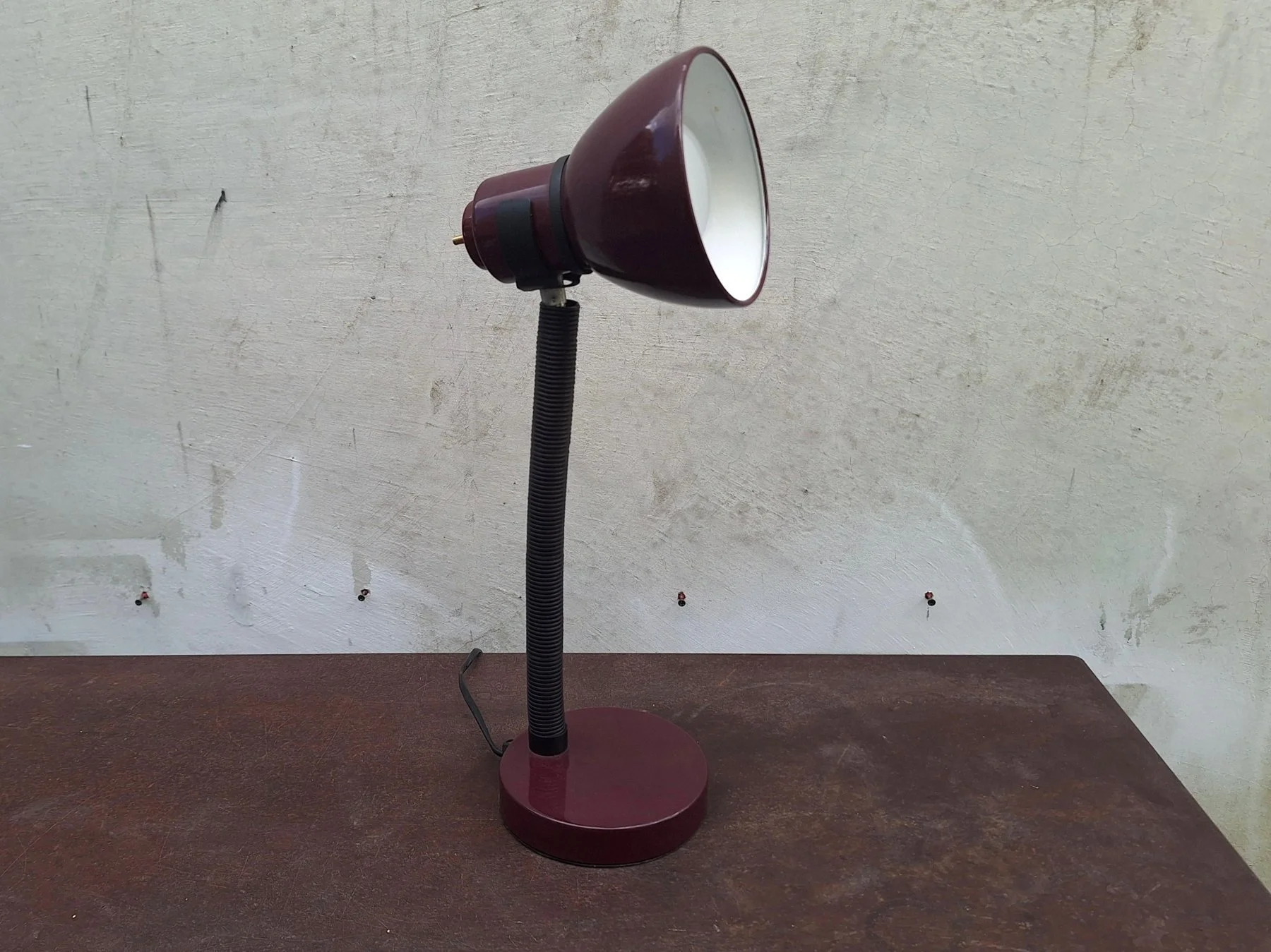 Vintage 1980s burgundy painted steel desk lamp with an adjustable gooseneck and directional shade photographed against a neutral concrete backdrop.