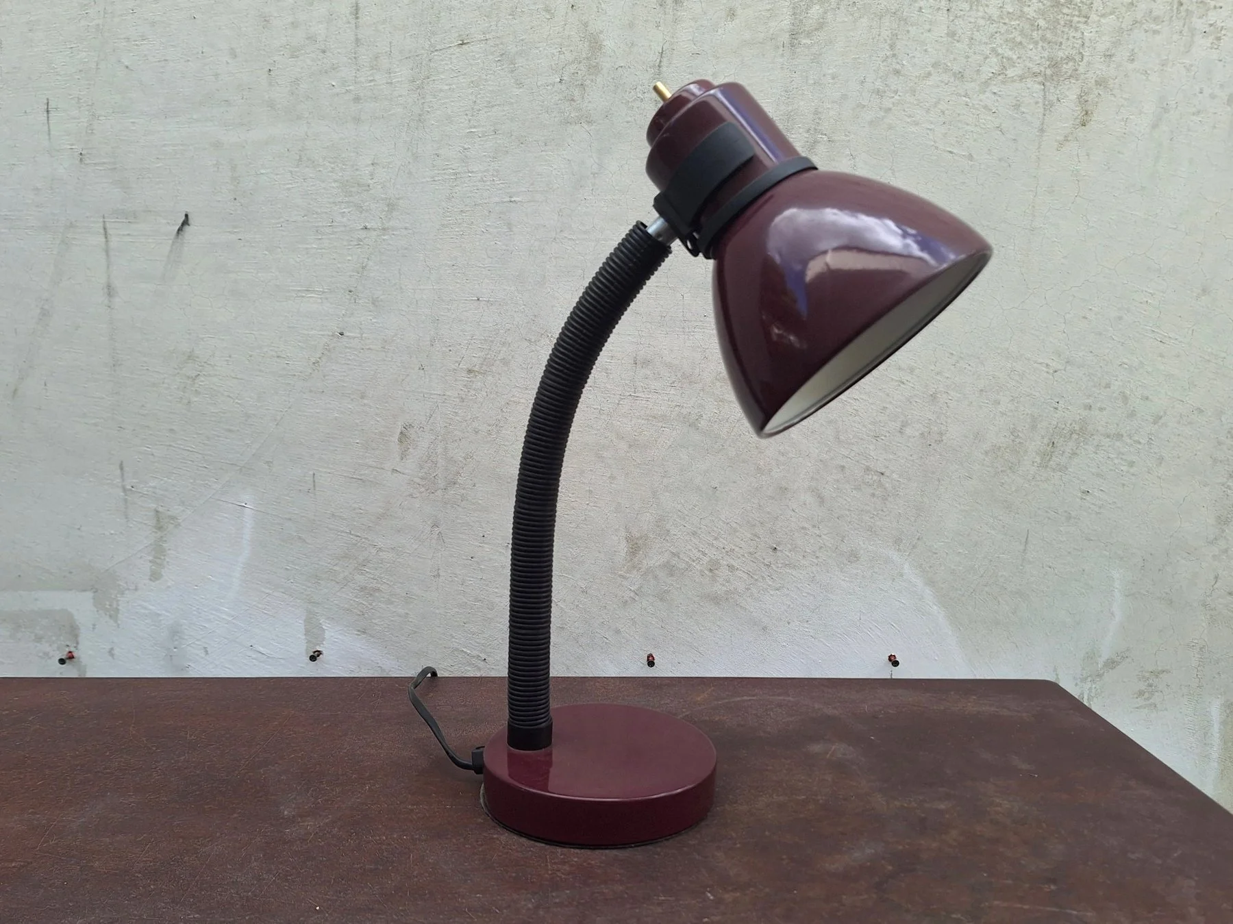 Vintage 1980s burgundy painted steel desk lamp with an adjustable gooseneck and directional shade photographed against a neutral concrete backdrop.