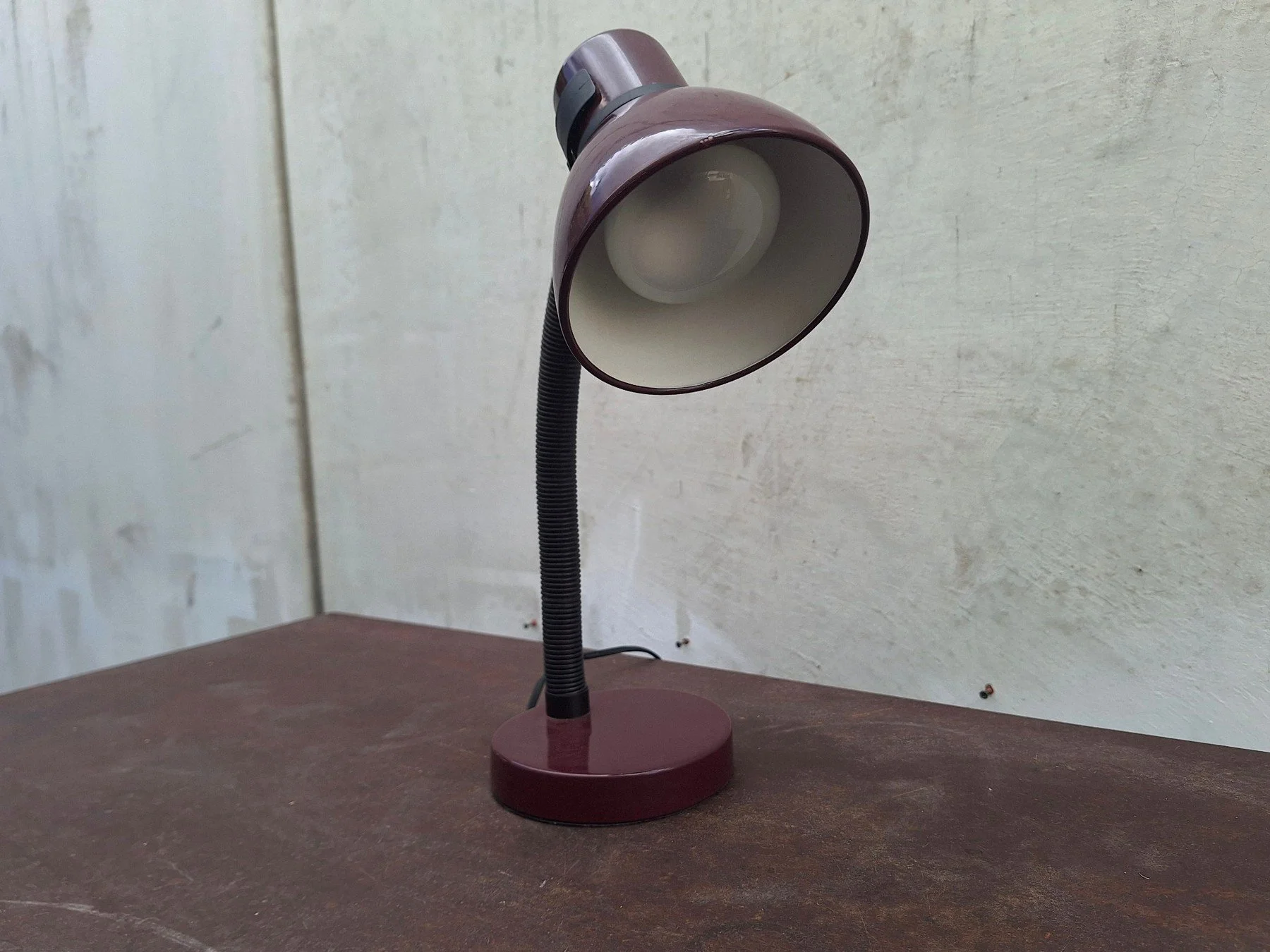 Vintage 1980s burgundy painted steel desk lamp with an adjustable gooseneck and directional shade photographed against a neutral concrete backdrop.