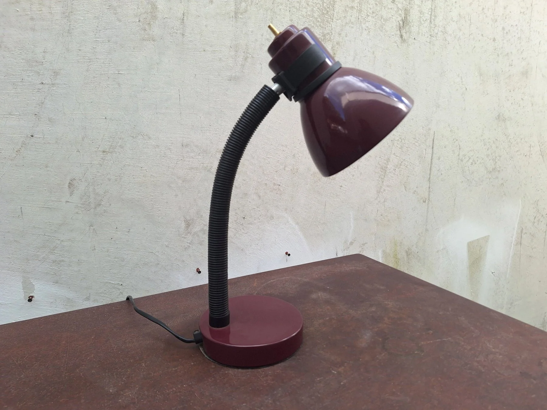 Vintage 1980s burgundy painted steel desk lamp with an adjustable gooseneck and directional shade photographed against a neutral concrete backdrop.