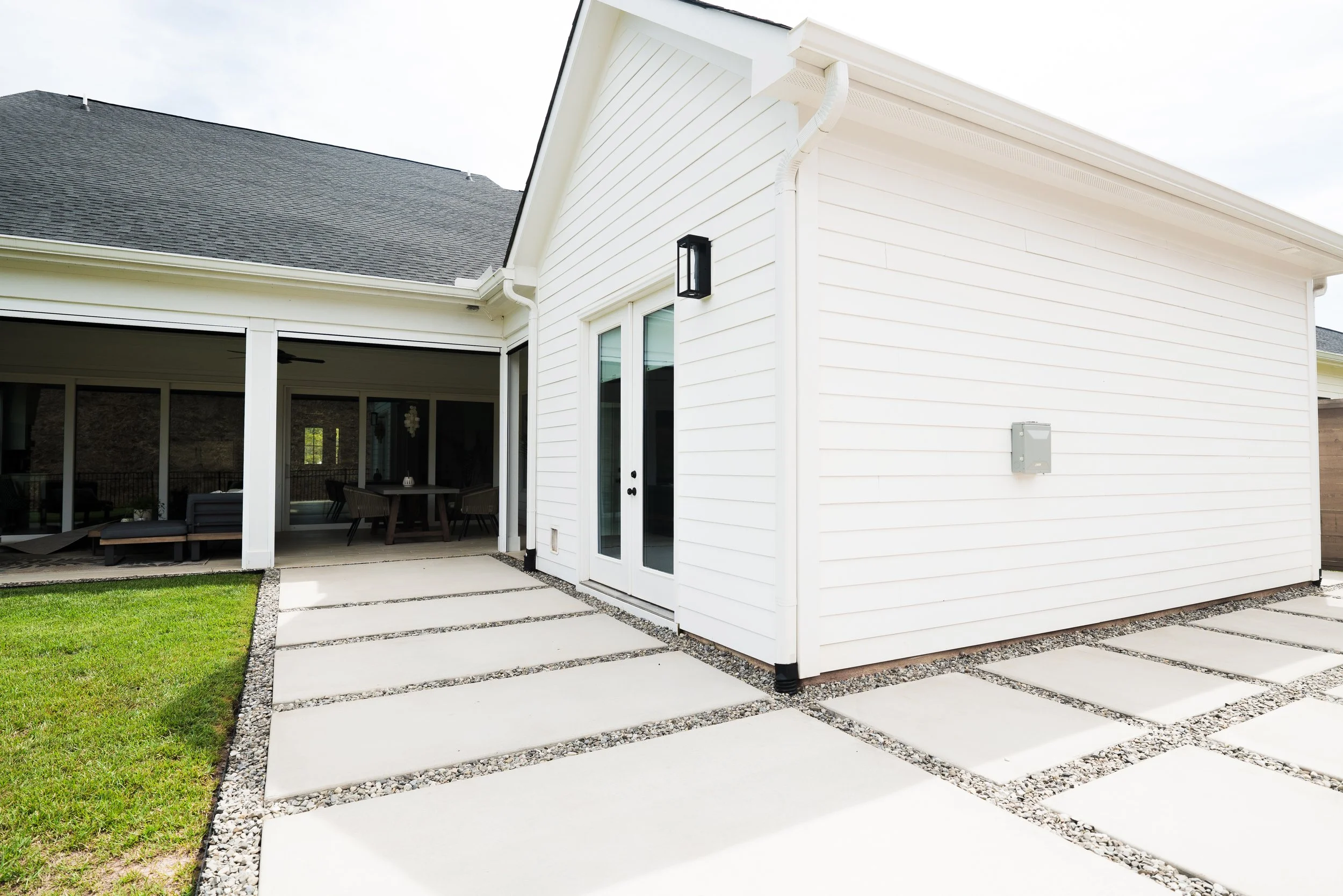 Backyard patio with concrete slabs separated by small gravel strips, adjacent to a house with white siding and glass double doors, with a covered patio area visible through the doors.