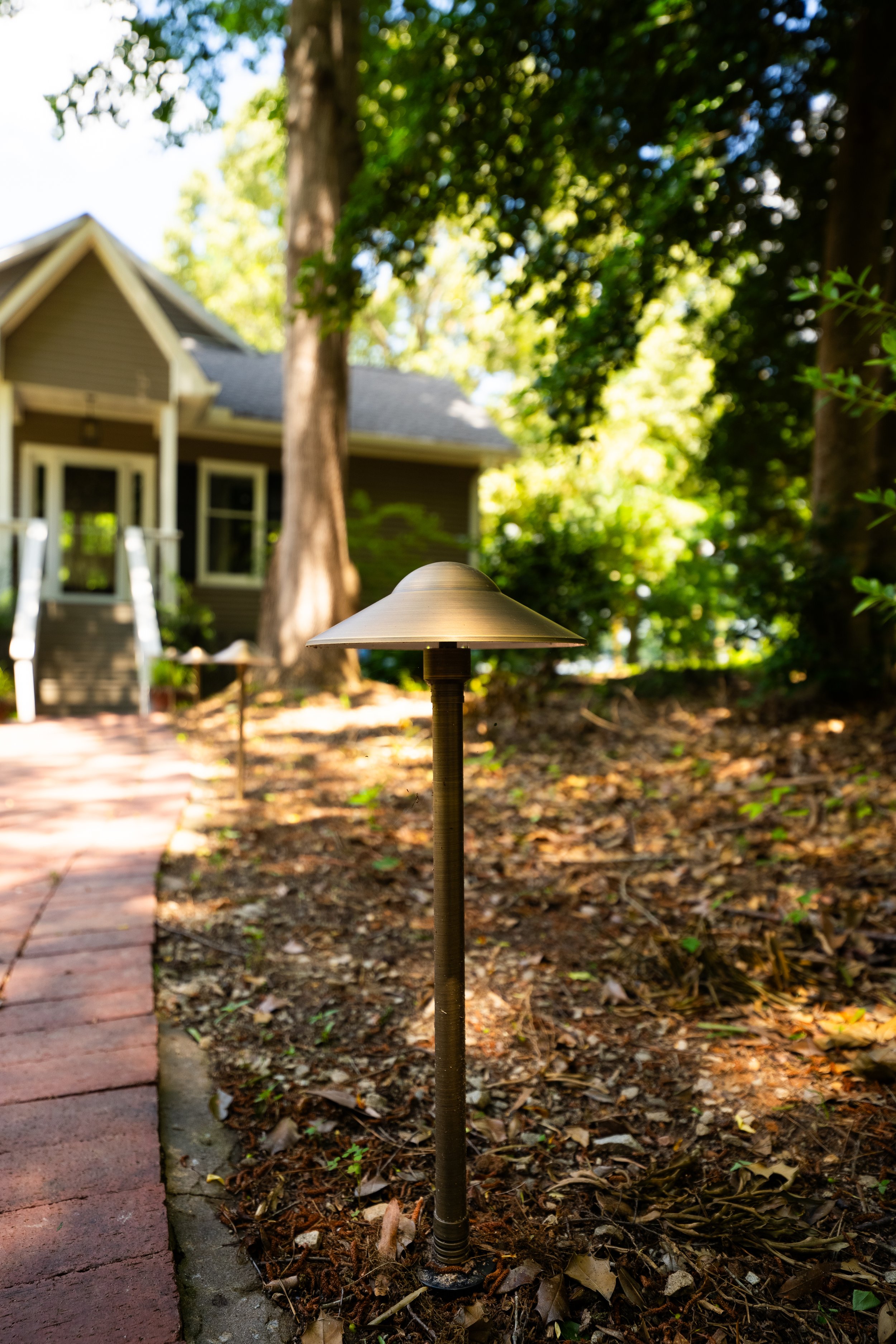 Close-up of a garden lamp post along a brick pathway leading to a house, with trees and greenery in the background.