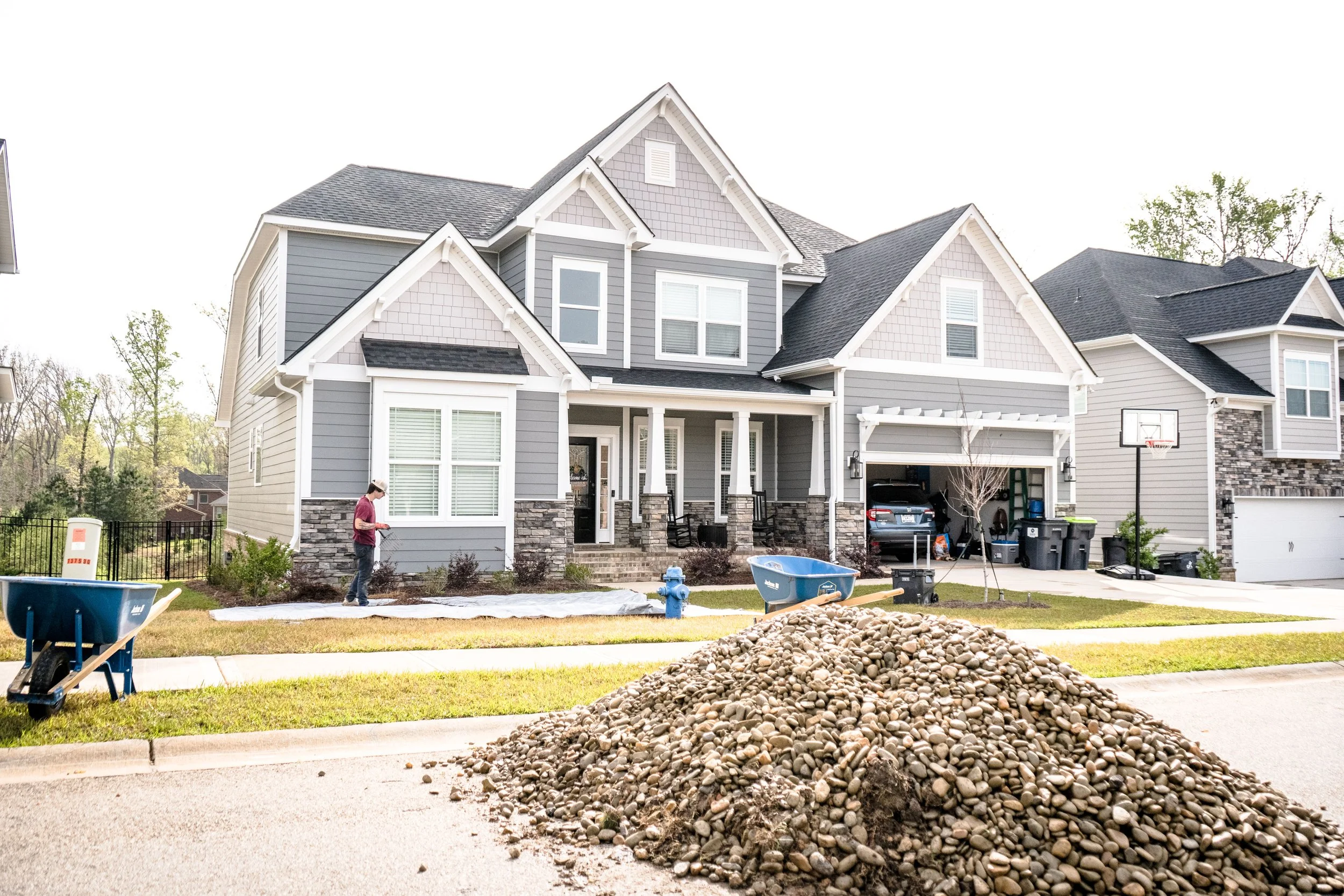 A multi-story house with gray siding and stone accents under renovation, with a large pile of rocks in the foreground, a worker on the lawn, wheelbarrows, and construction tools and supplies nearby.