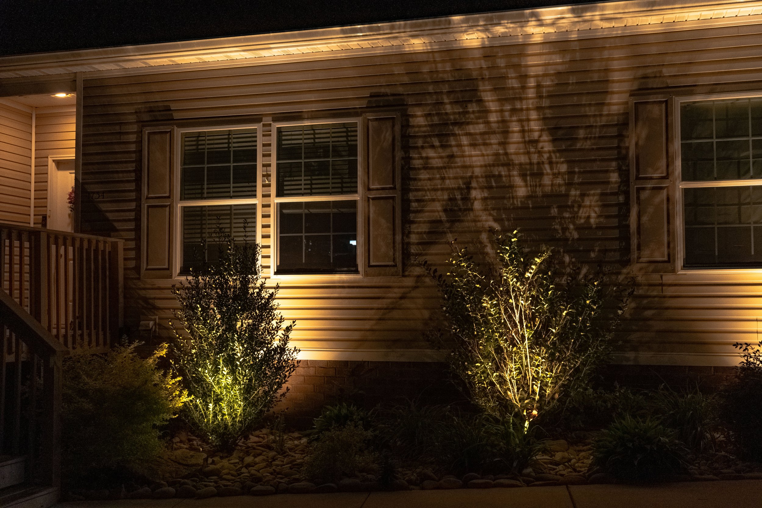 Nighttime view of the exterior of a house with illuminated bushes and windows, showing a porch area on the left side.