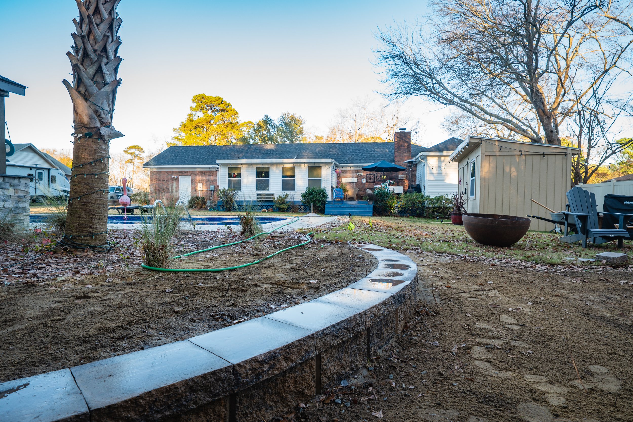 A backyard with a brick house and a patio area, a palm tree wrapped with string lights, garden bed with a stone border, and outdoor seating including a Adirondack chair and a fire pit, during late afternoon.