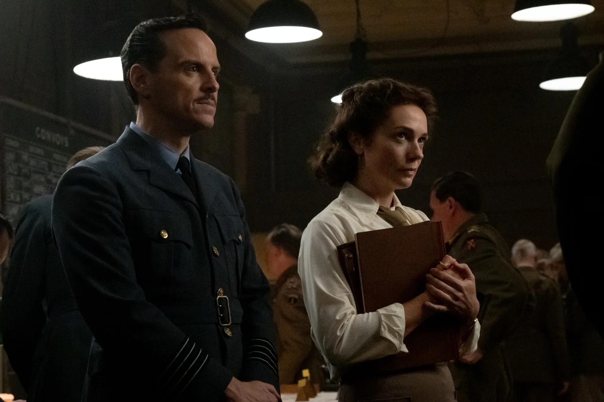 A weather officer and a military secretary stand among Allied commanders inside a crowded wartime briefing room in WWII drama Pressure.