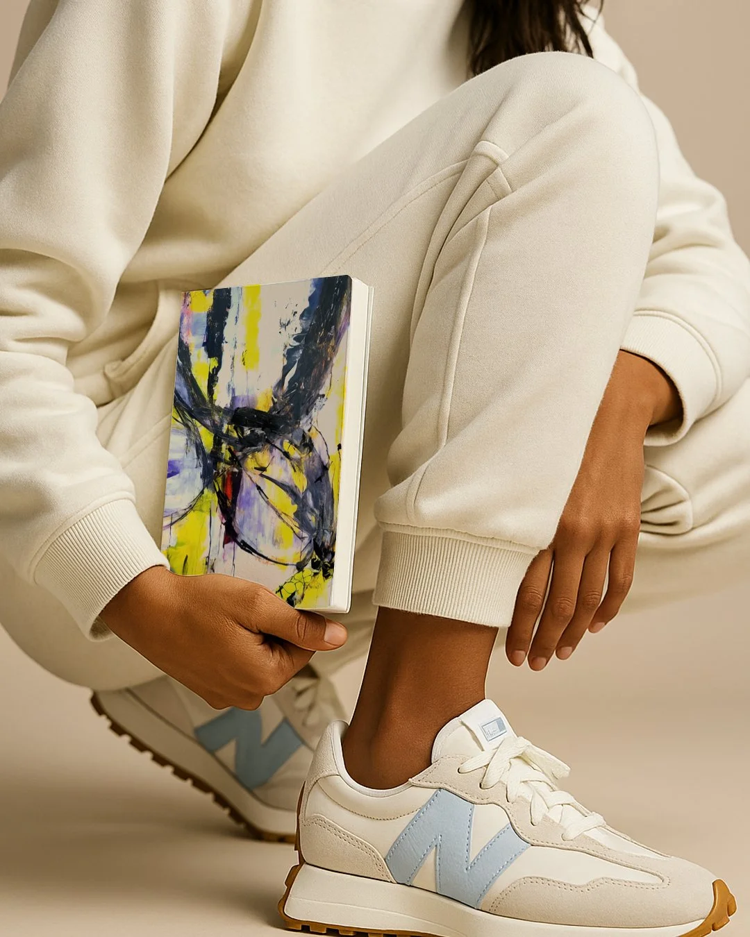 Person wearing white sneakers with light blue accents, beige sweatpants, and a cream hoodie, holding a colorful abstract art book.
