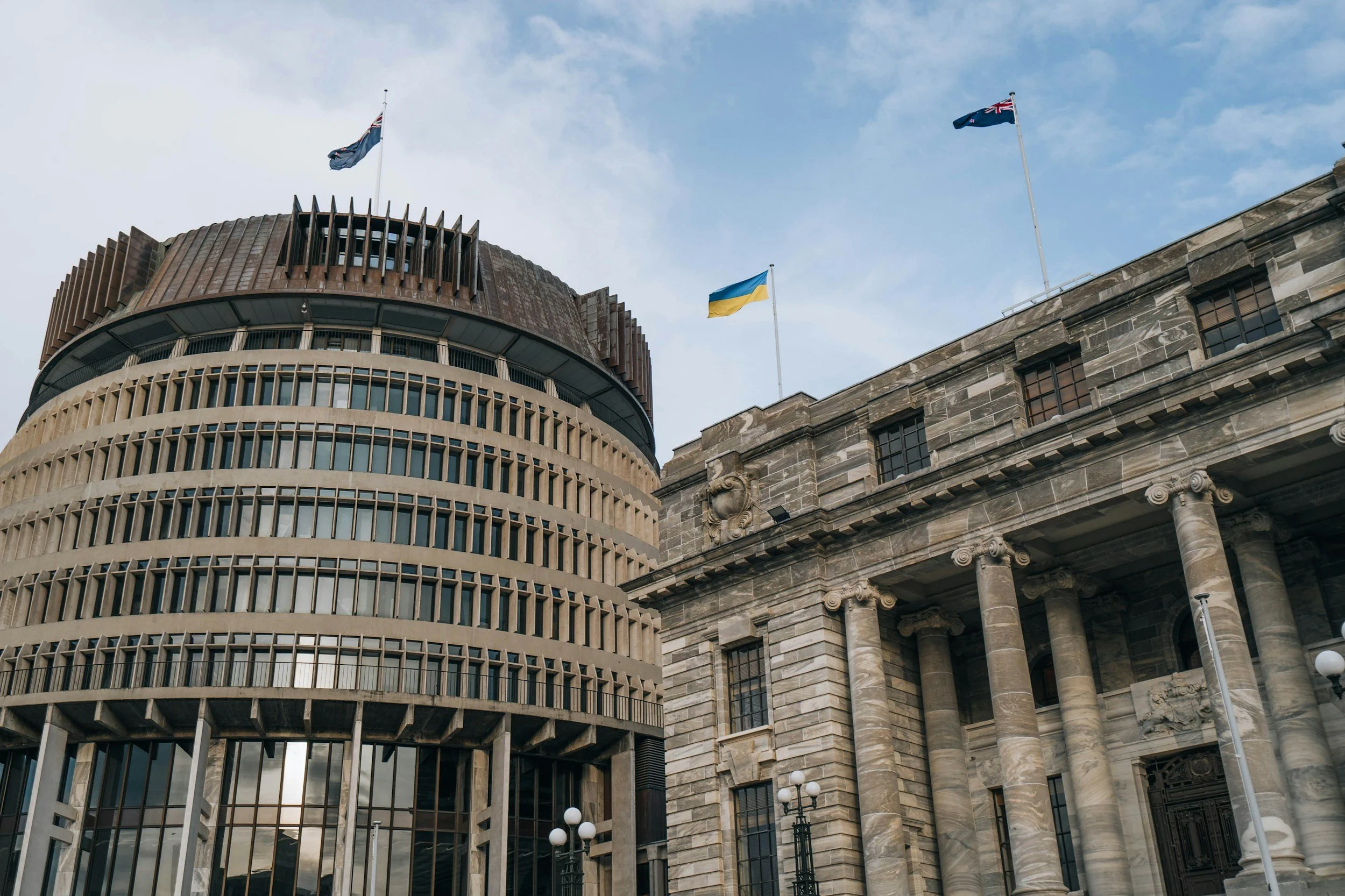 A modern, circular government building with vertical glass windows and a government building with classical columns and a carved frieze, both flying flags, under a partly cloudy sky.