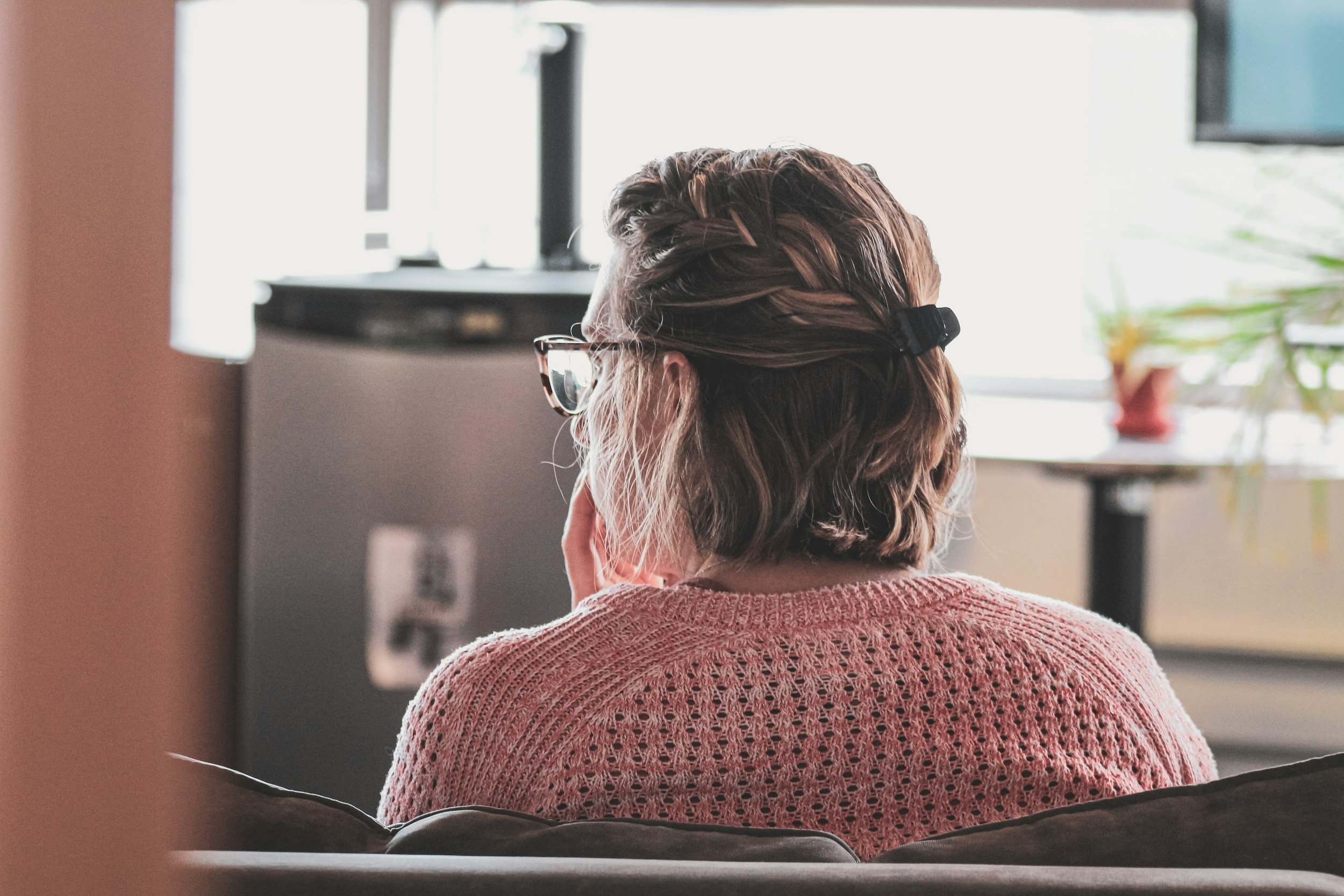 A woman with short brown hair wearing glasses and a pink knitted sweater, sitting on a sofa, facing away from the camera, looking at a television in a room with natural light.