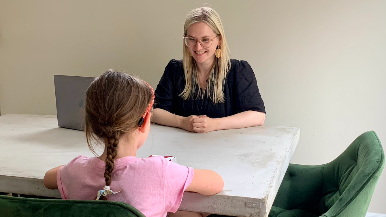 A woman with blonde hair and glasses smiling at a girl with brown hair in a braid, sitting at a table. The woman is wearing a black shirt and large earrings, and the girl is wearing a pink shirt. There is a gray laptop and a small white box on the table, and a green chair is visible.
