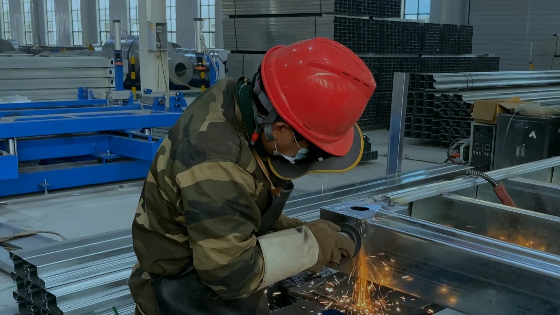 Worker wearing a red safety helmet and camouflage jacket welding metal pipes in an industrial warehouse.