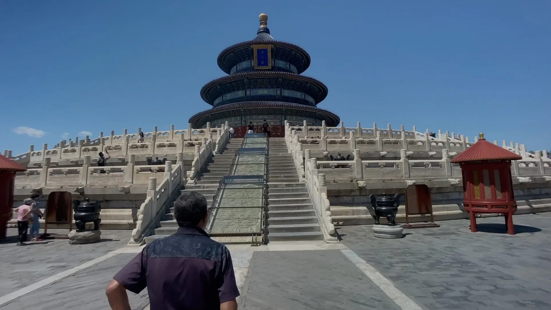 A large, multi-tiered pagoda-style building with white marble stairs leading up to it. The building has a dark, ornate roof and is situated under a clear blue sky. Visitors are walking around the area.