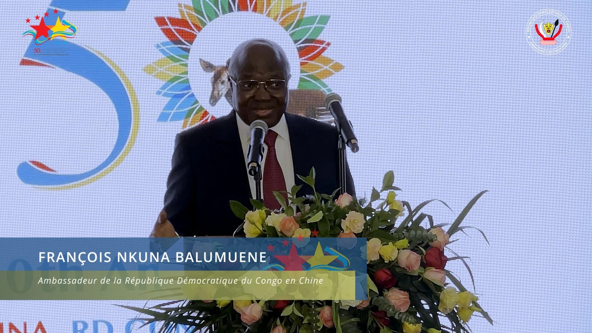 A man in a suit and red tie speaking at a podium with a bouquet of flowers, in front of a colorful backdrop featuring a star and a horse, with text overlay about François Nkuna Balumuene, the Democratic Republic of Congo ambassador to China.