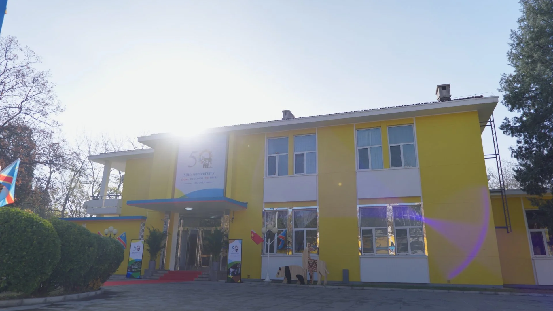 A yellow two-story building with white accents, large windows, and a small balcony on the second floor. There are banners celebrating a 50th anniversary, flags, and decorative lion statues outside. The sun is shining brightly, creating lens flare, an